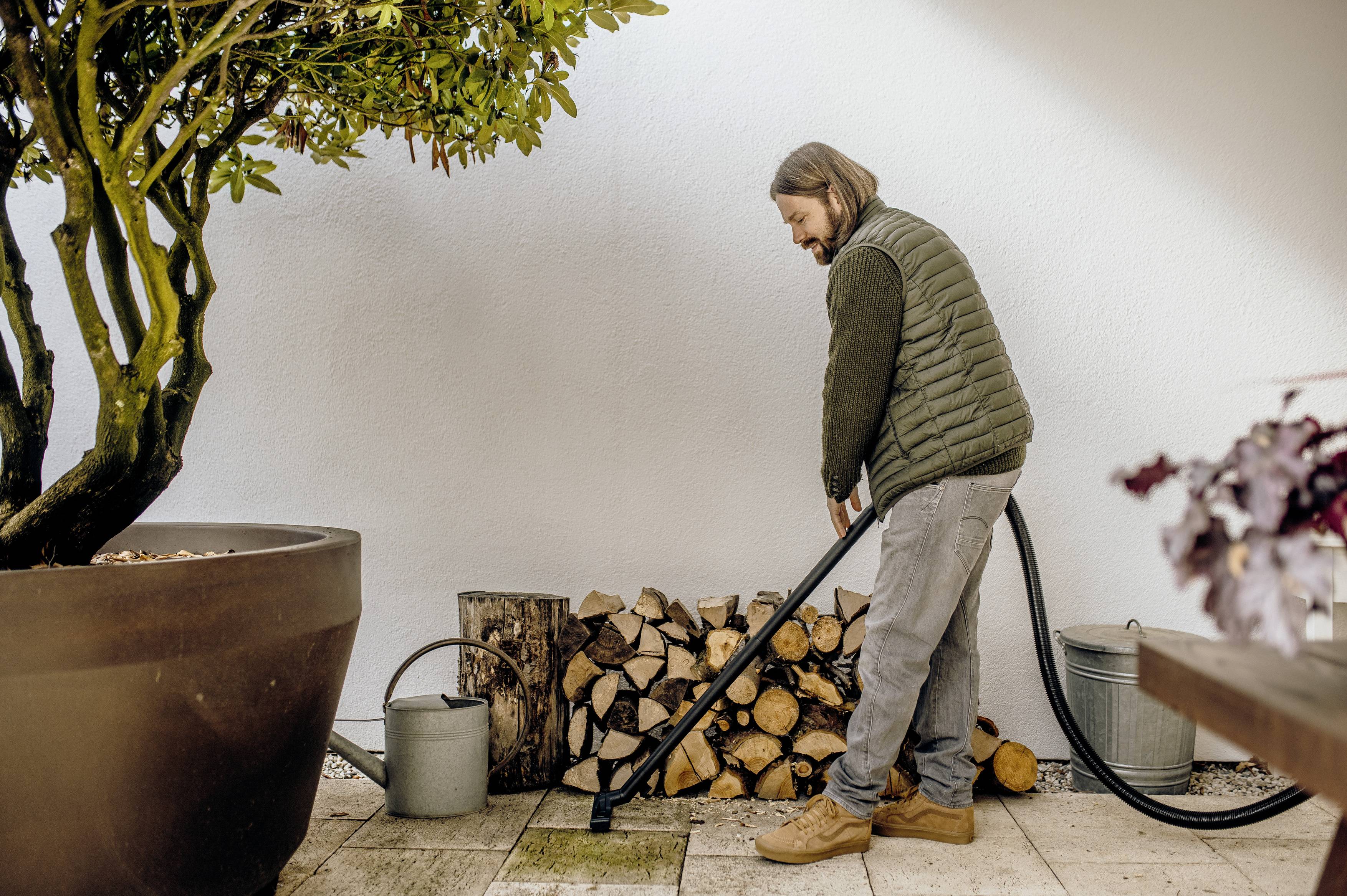 Eine Person gießt Pflanzen mit einem Schlauch in einem Garten. Es gibt gestapelte Holzscheite und Gießkannen in der Nähe.