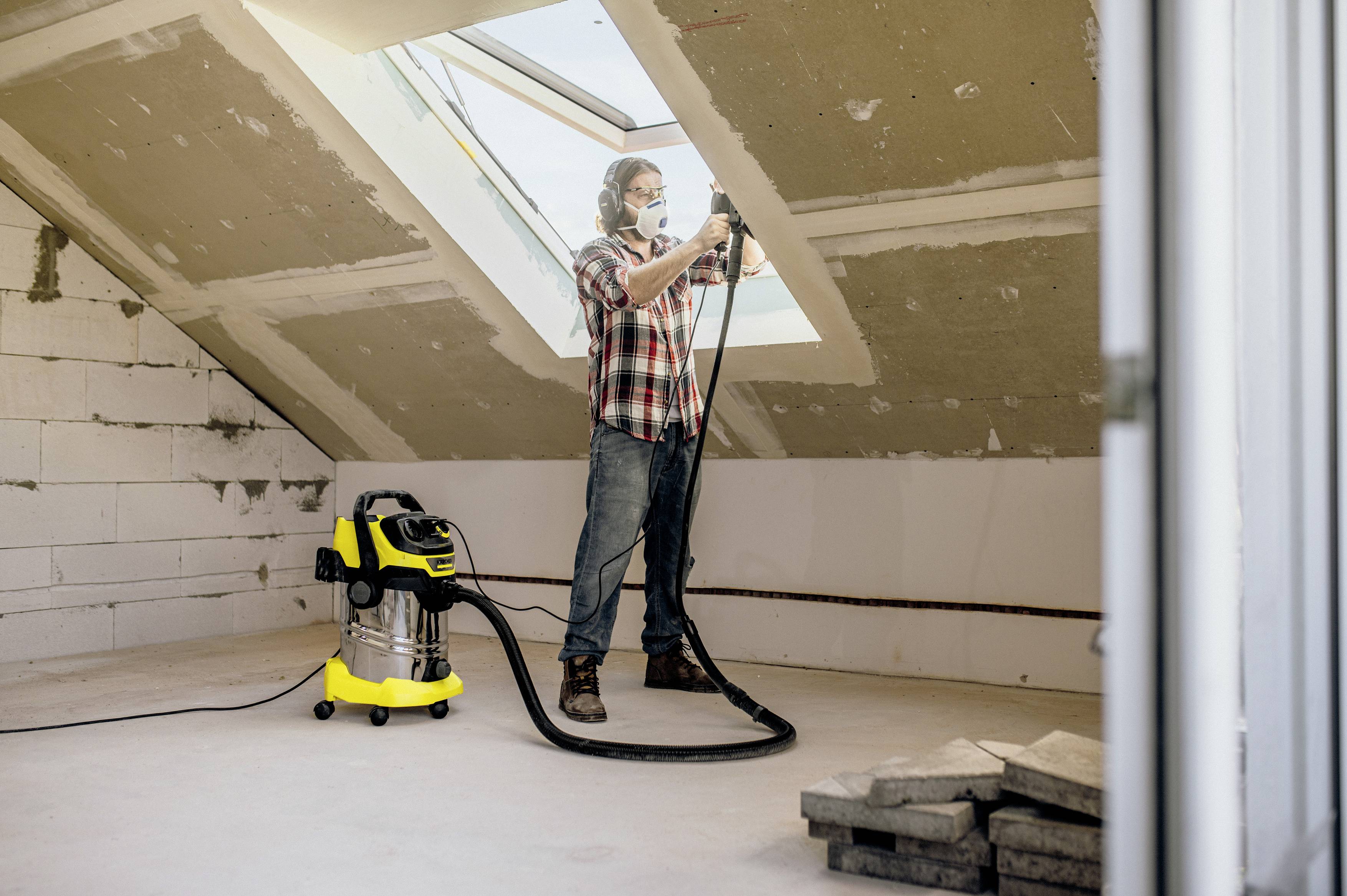 Eine Person reinigt mit einem Industriestaubsauger ein Dachgeschosszimmer, das durch ein großes Dachfenster beleuchtet wird.