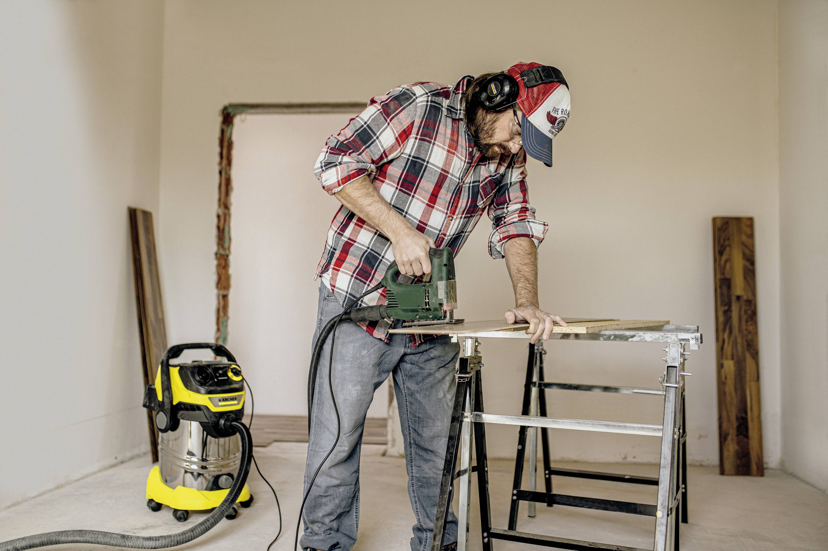 Ein Mann trägt ein Holzfällerhemd und eine Mütze, arbeitet mit einer elektrischen Stichsäge an einem Stück Holz. Im Hintergrund steht ein Staubsauger.
