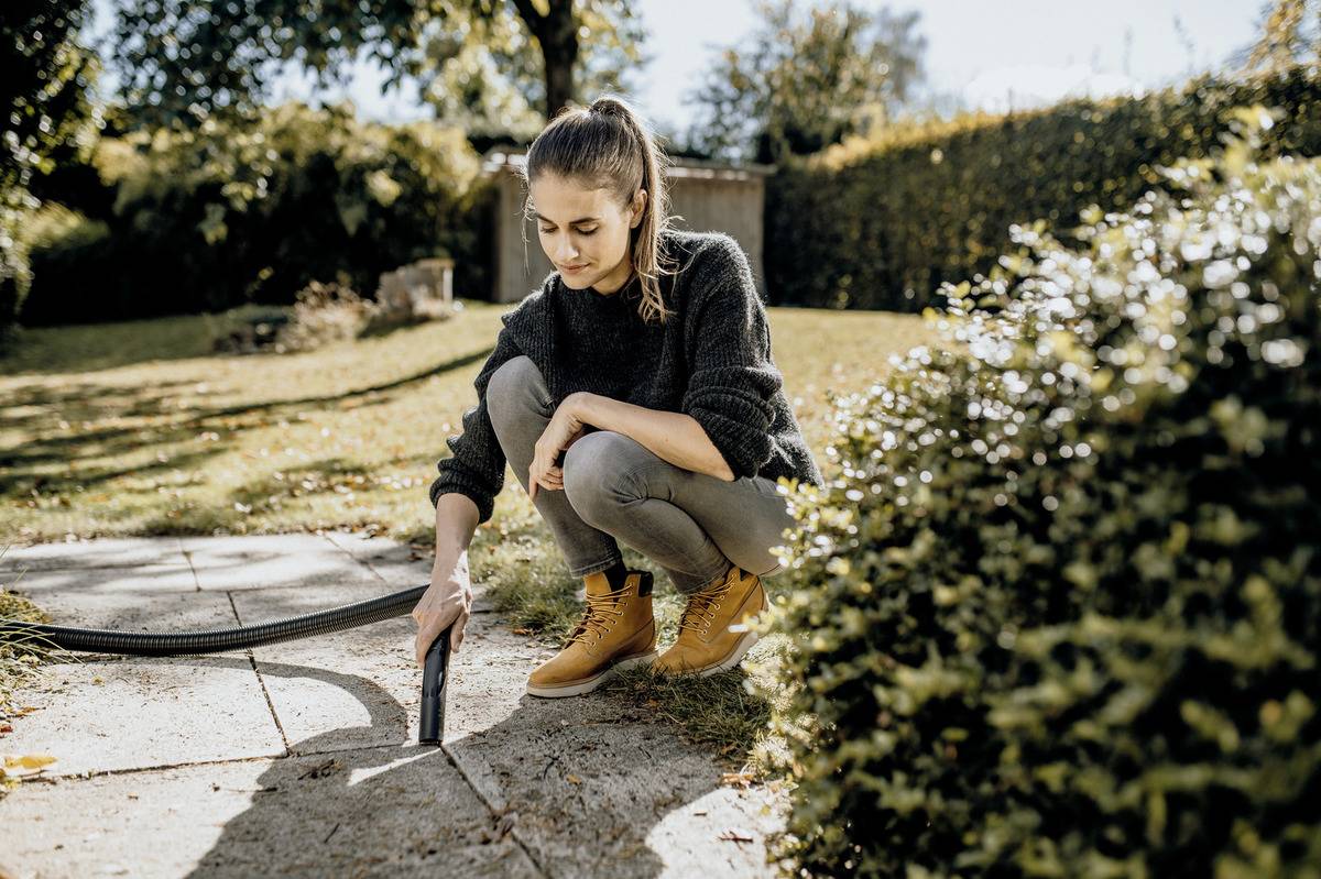 Eine Person kniet auf einem Gartenweg und verwendet einen Staubsauger, um zwischen den Pflastersteinen zu reinigen. Von Sonnenlicht beschienene Bäume und Sträucher umgeben den ordentlichen Garten.