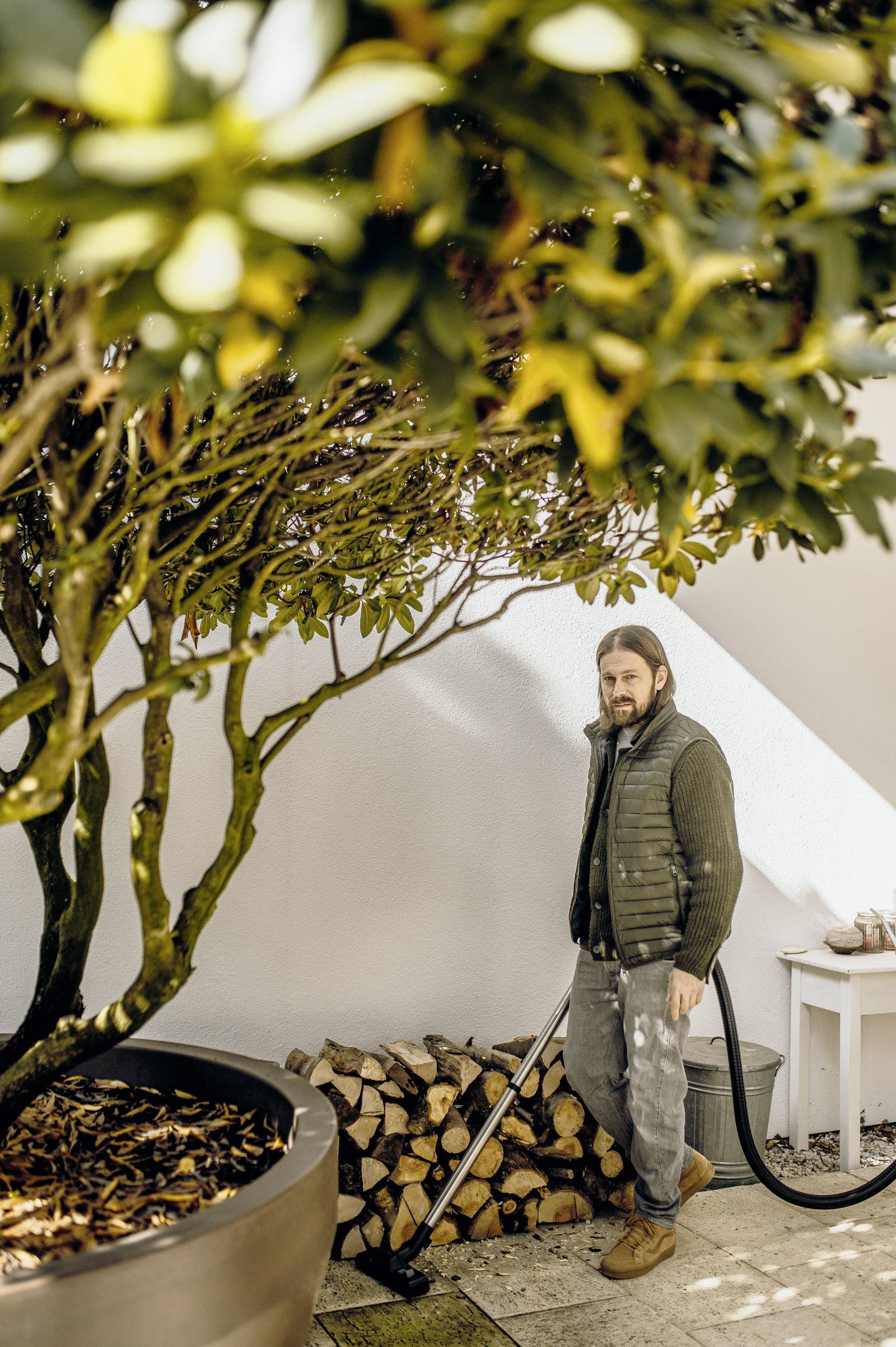 Eine Person, die neben einem Stapel Brennholz steht und einen Staubsaugerschlauch hält, unter einem Baum in einer Hinterhofumgebung mit weißer Wandkulisse.