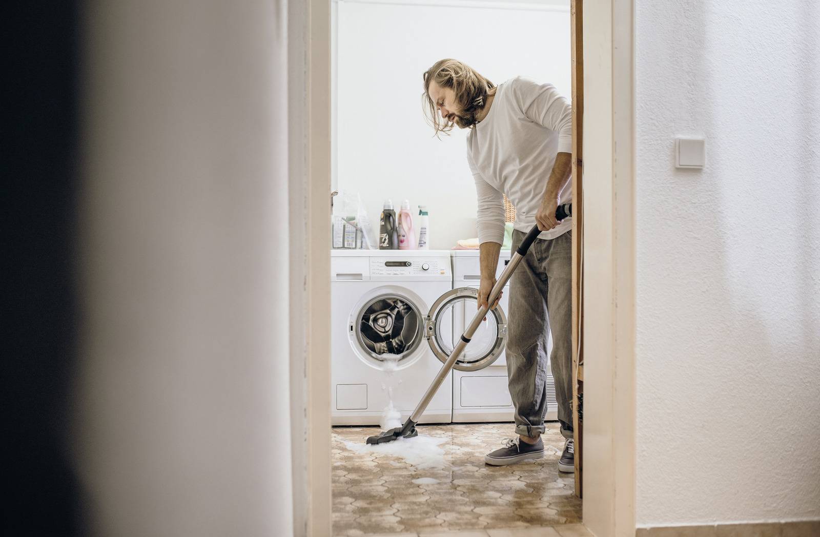 Eine Person saugt einen Teppich in einem Waschraum, während eine Waschmaschine, ein Trockner und Reinigungsutensilien im Hintergrund zu sehen sind.