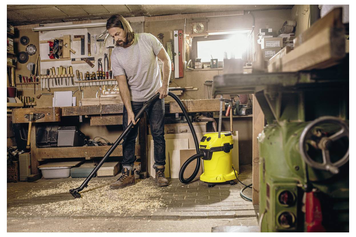 Eine Person in einer Werkstatt, die einen Staubsauger benutzt, um Holzspäne vom Boden zu reinigen. Verschiedene Werkzeuge und eine vollständig bestückte Werkbank sind zu sehen.