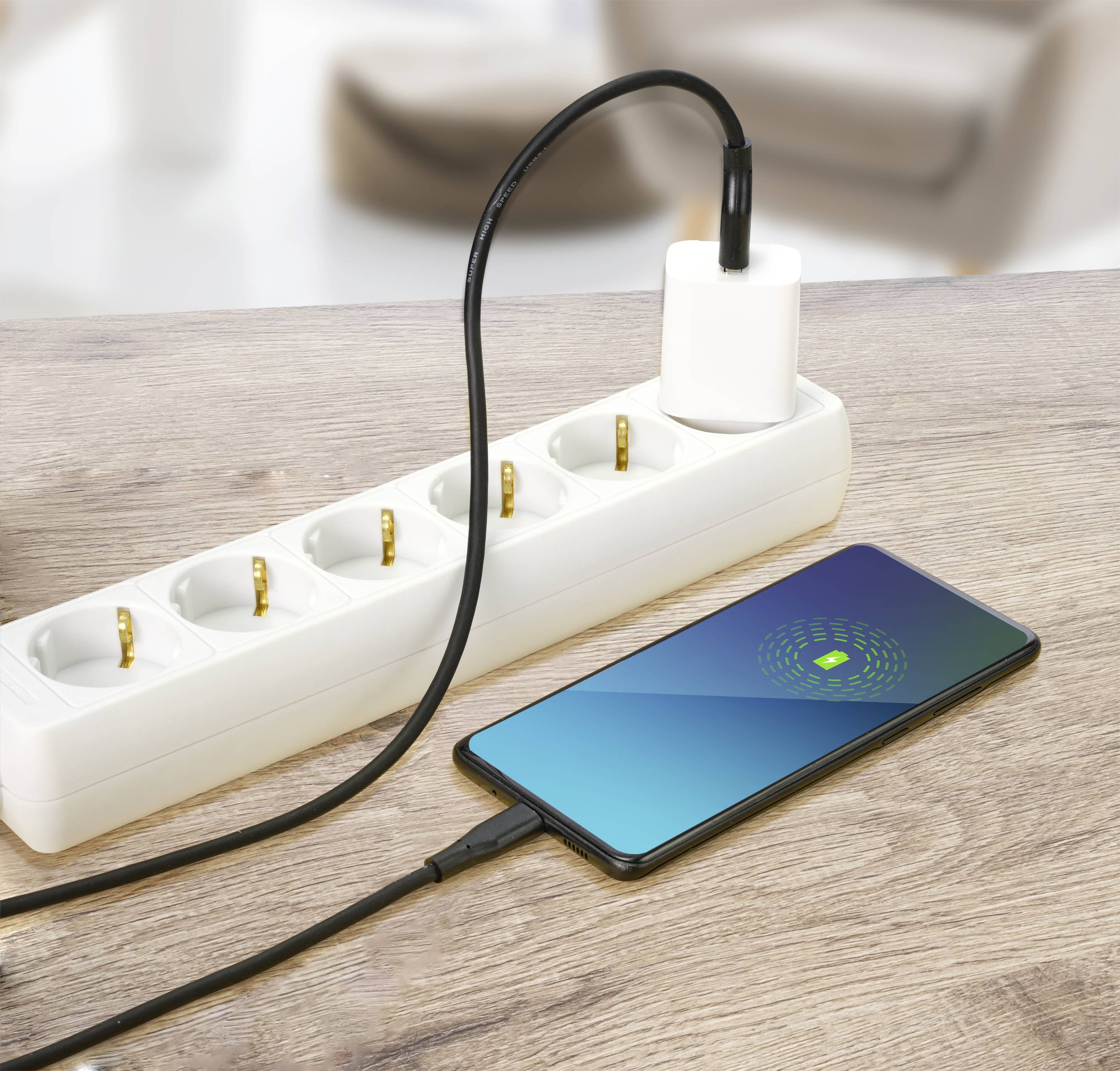 Smartphone auf Tisch wird über Verlängerungskabel geladen, das in Steckdose eingesteckt ist. Fokus liegt auf Ladevorgang.