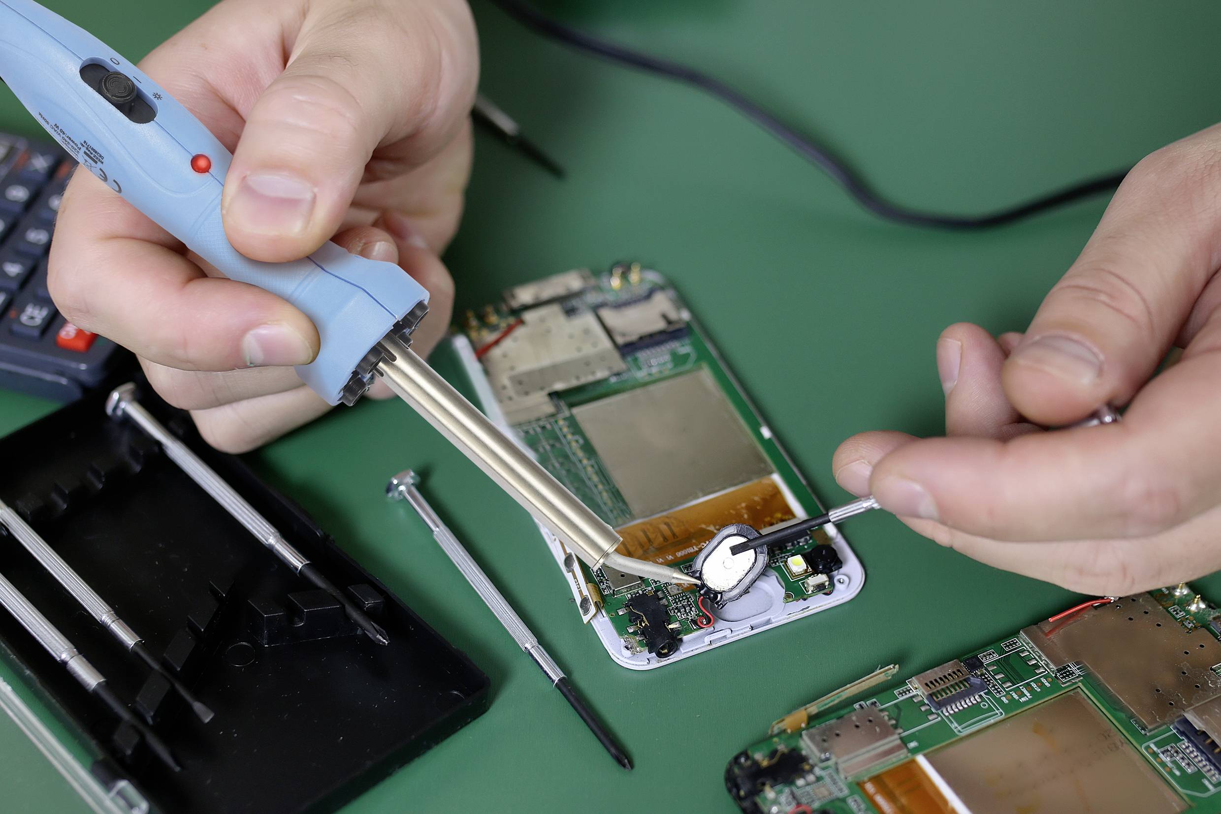 Eine Person repariert eine geöffnete Handyplatine mit einem Lötkolben. Schraubendreher und weitere Werkzeuge liegen daneben.