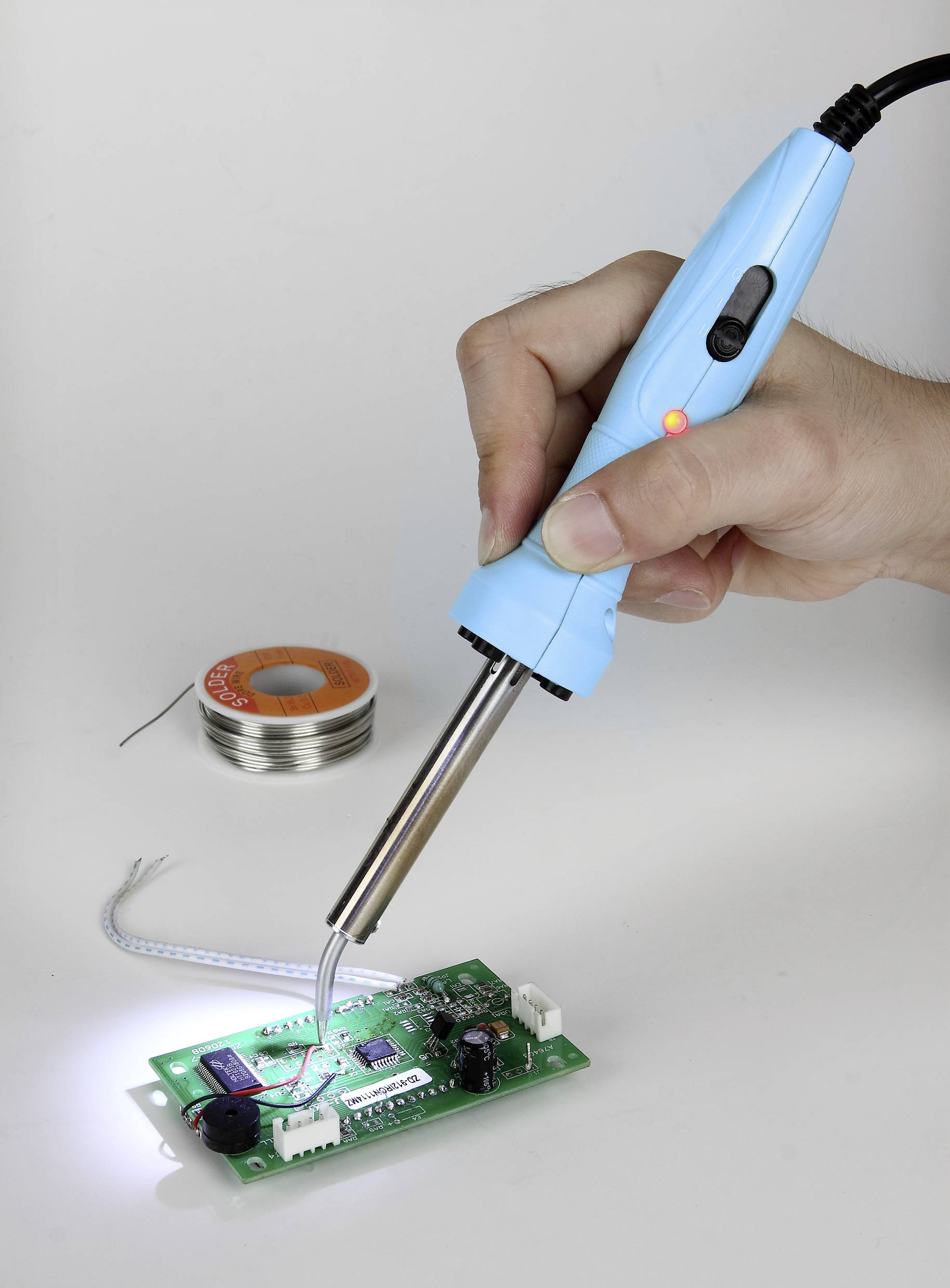 Person hält einen Lötkolben und lötet Drähte auf eine grüne Leiterplatte. Im Hintergrund ist eine Rolle Lötzinn zu sehen.