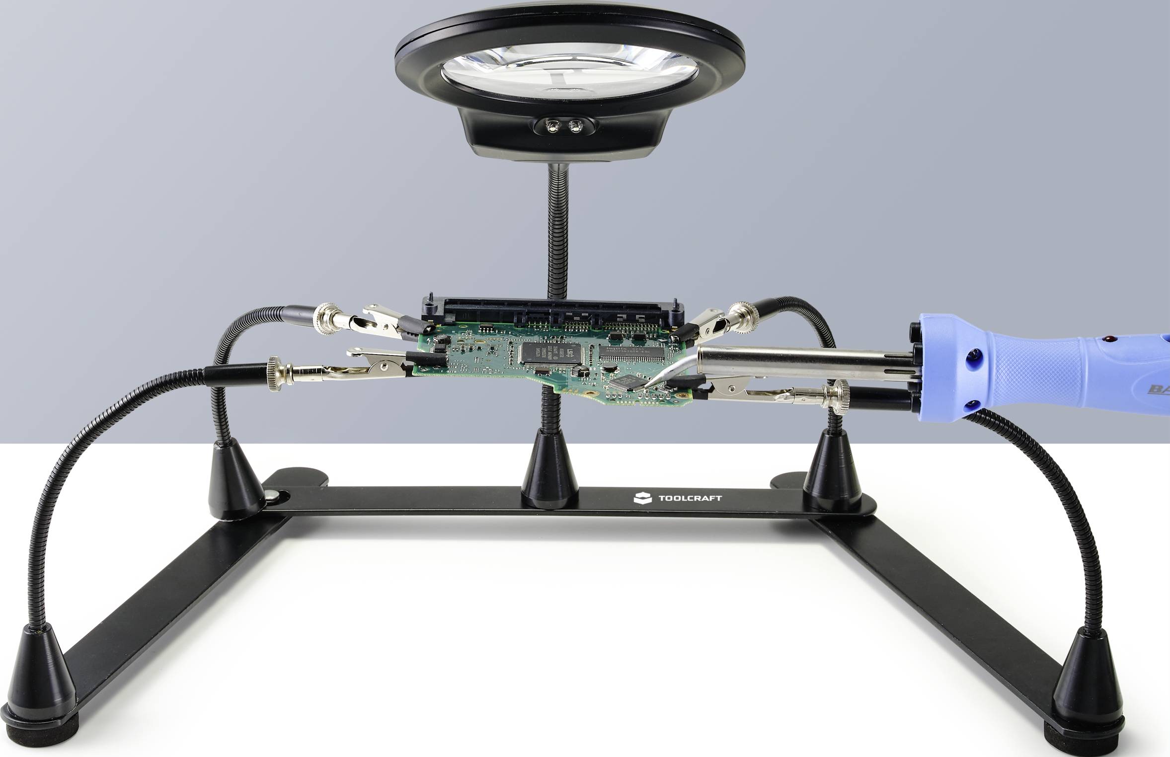 Bild zeigt eine Lötstation mit Platine unter einer Vergrößerungslinse. Ein Lötkolben berührt die Platine, bereit zur Bearbeitung.