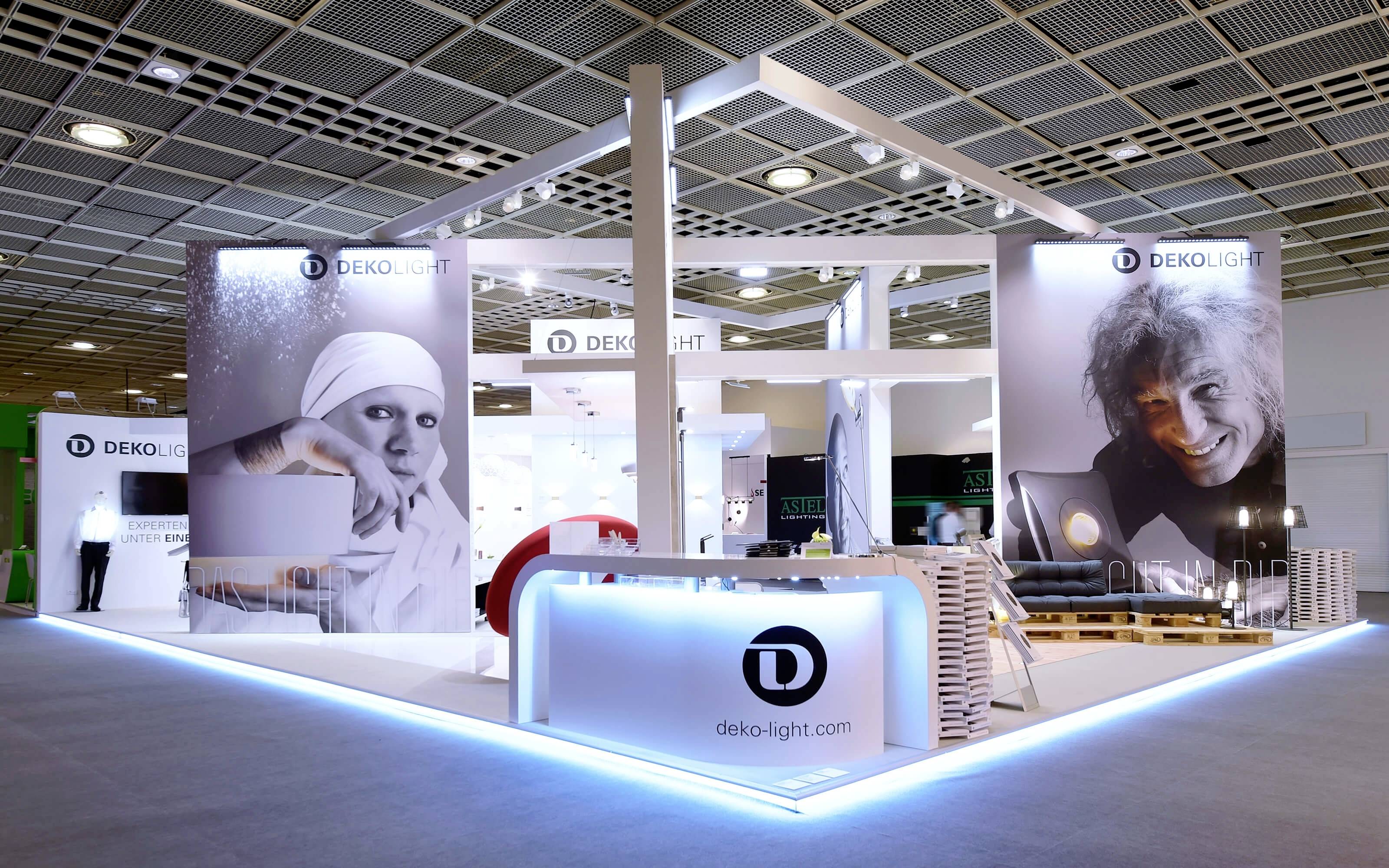 Messestand von DEKO-LIGHT mit großen Porträtbildern, Logos und einem beleuchteten Tresen, präsentiert auf einer Messe in einer großen Halle.