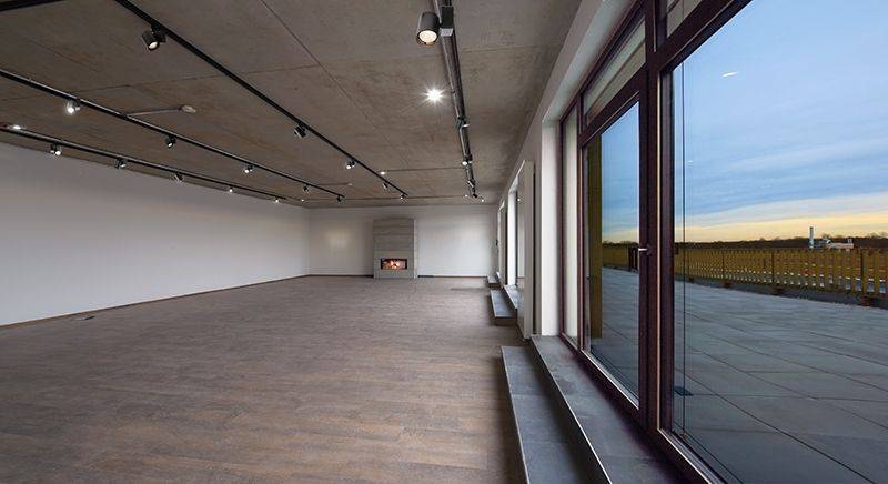 Ein leerer Raum mit Holzboden, großen Fenstern und einem Kamin an der Wand. Außenansicht zeigt eine Terrasse und einen weiten Himmel.