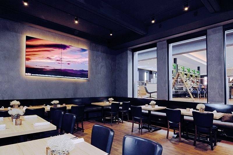 Ein modernes Restaurant mit Holzböden, schwarzen Stühlen und Tischen. Ein großes Gemälde zeigt einen farbenfrohen Sonnenuntergang an der Wand.