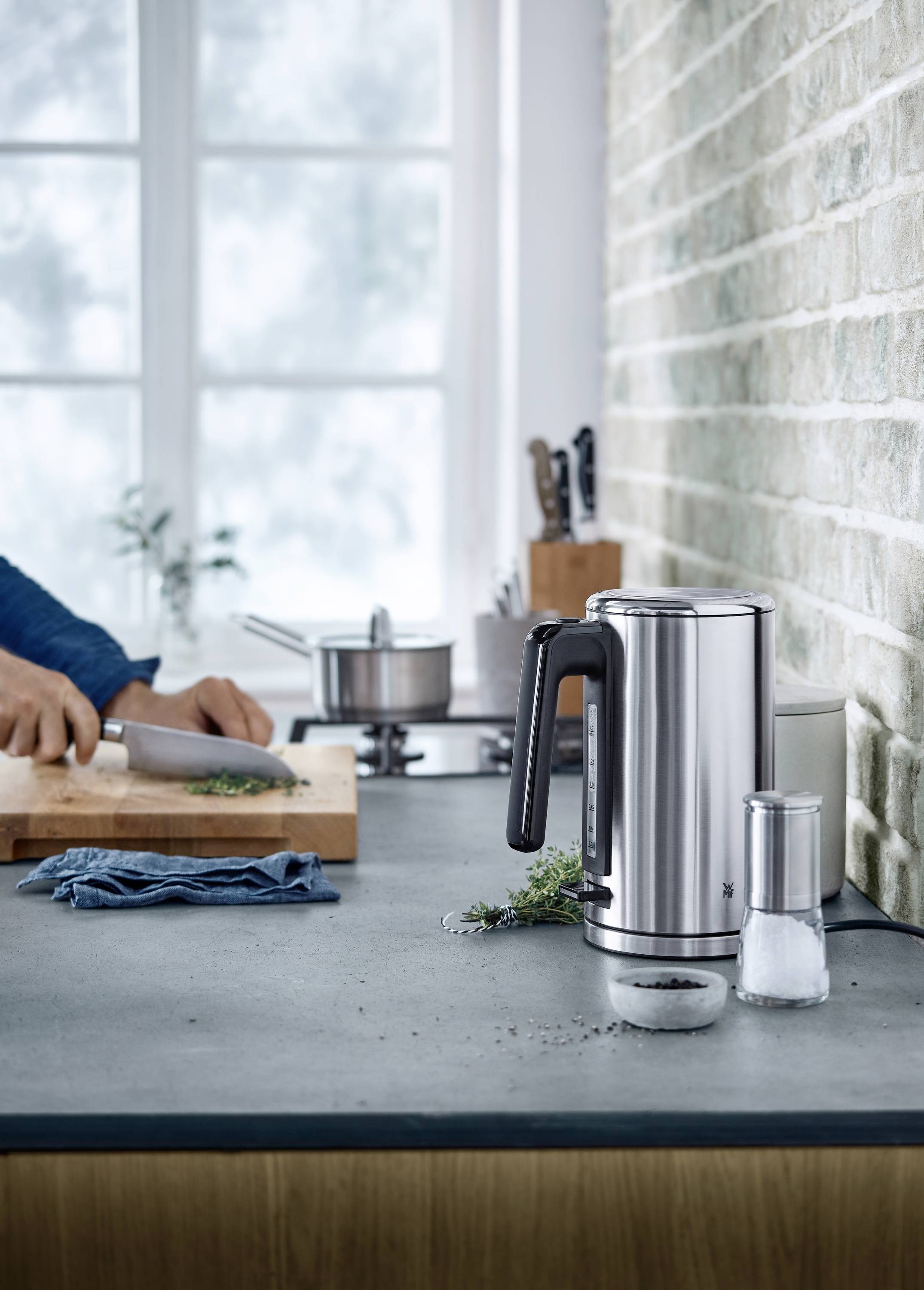 Eine Küche mit einem Wasserkocher und einer Gewürzmühle im Vordergrund. Im Hintergrund schneidet jemand Kräuter neben einem Kochtopf.