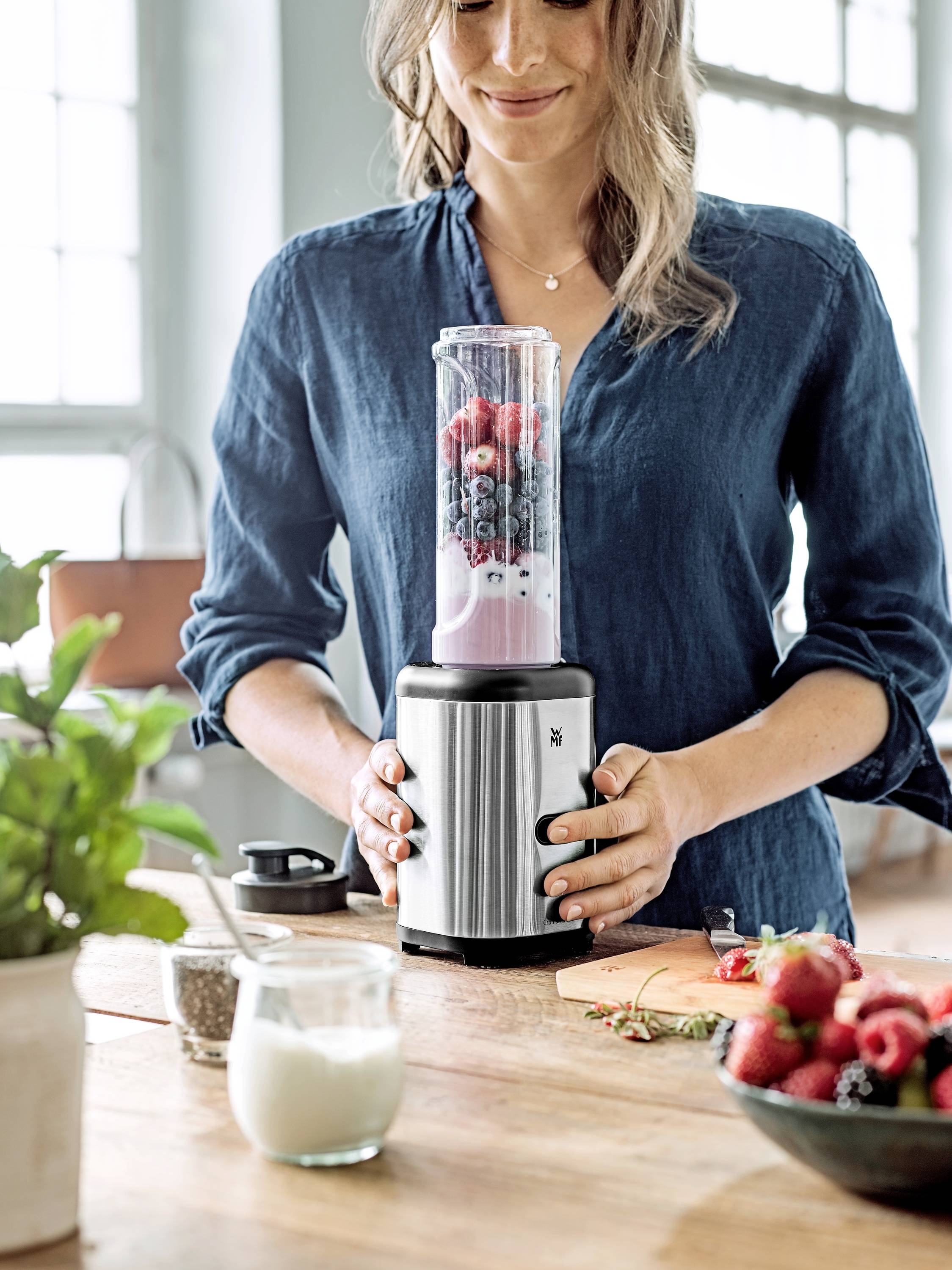 Frau in blauer Bluse benutzt Mixer, um Obst-Smoothie zu machen. Frische Beeren und Joghurt auf dem Tisch. Küche im Hintergrund.