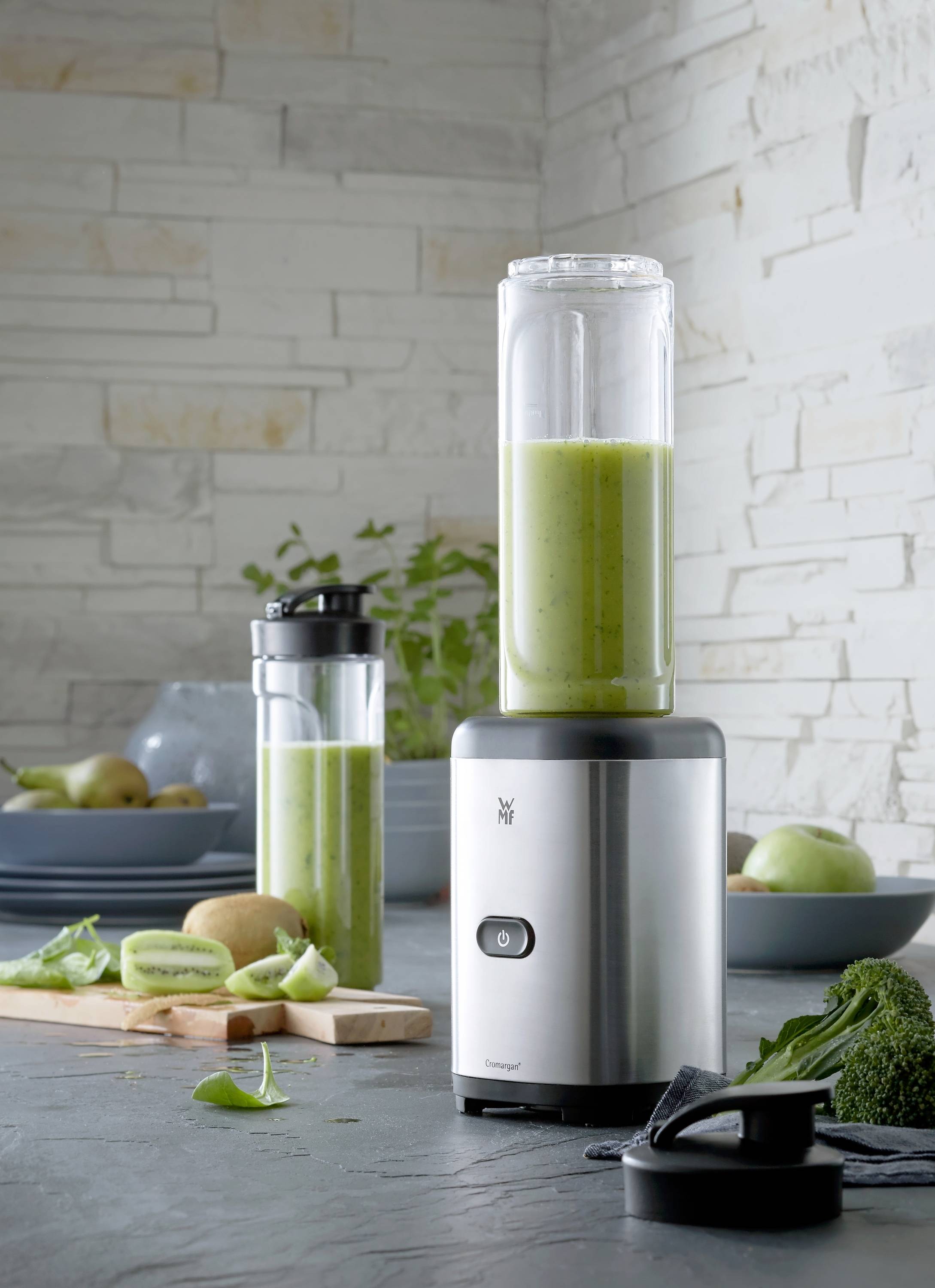Ein silberner Standmixer mit einem Glas grünen Smoothies steht auf einer Küchentheke, umgeben von Kiwi und Spinat. Im Hintergrund ein zweites Glas.