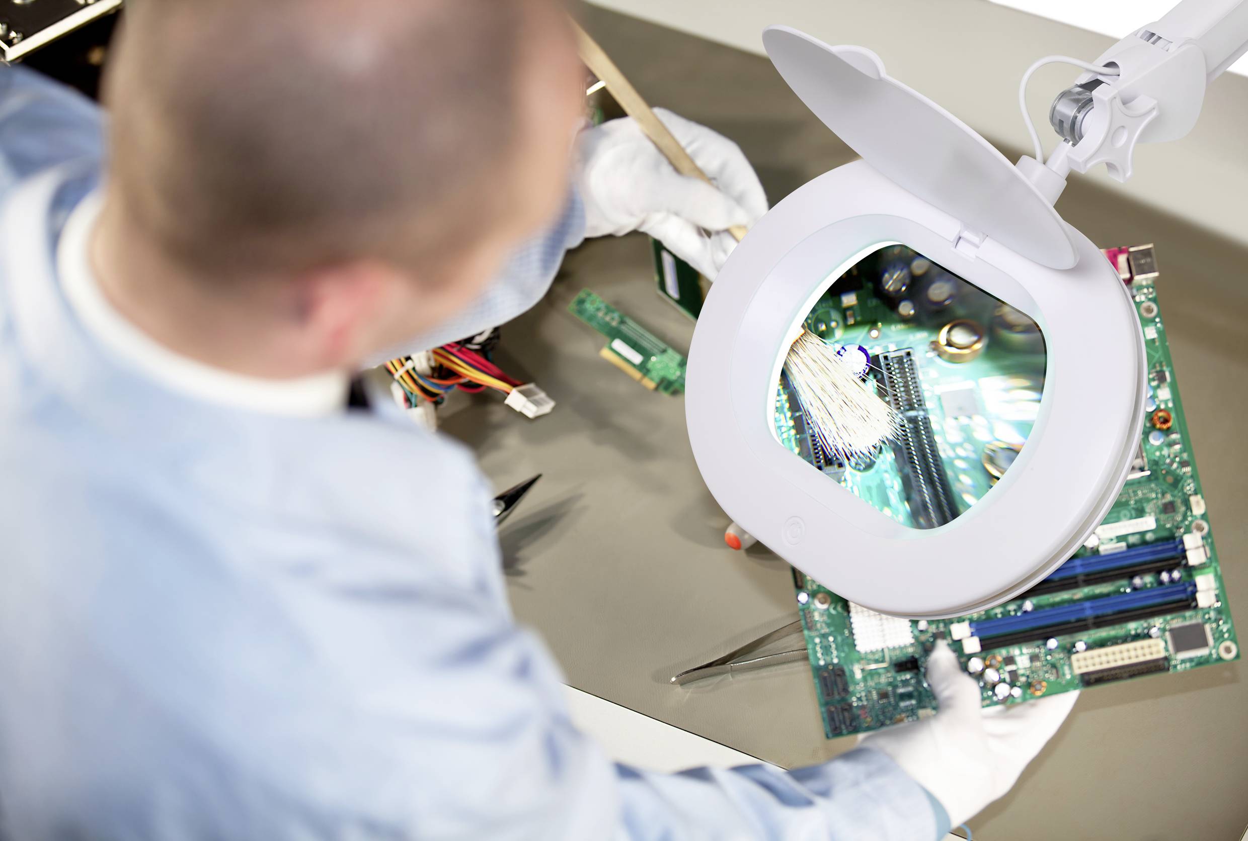 Eine Person in Schutzkleidung überprüft eine Computerplatine mit einer Lupenlampe, um die elektronischen Komponenten genauer zu inspizieren.