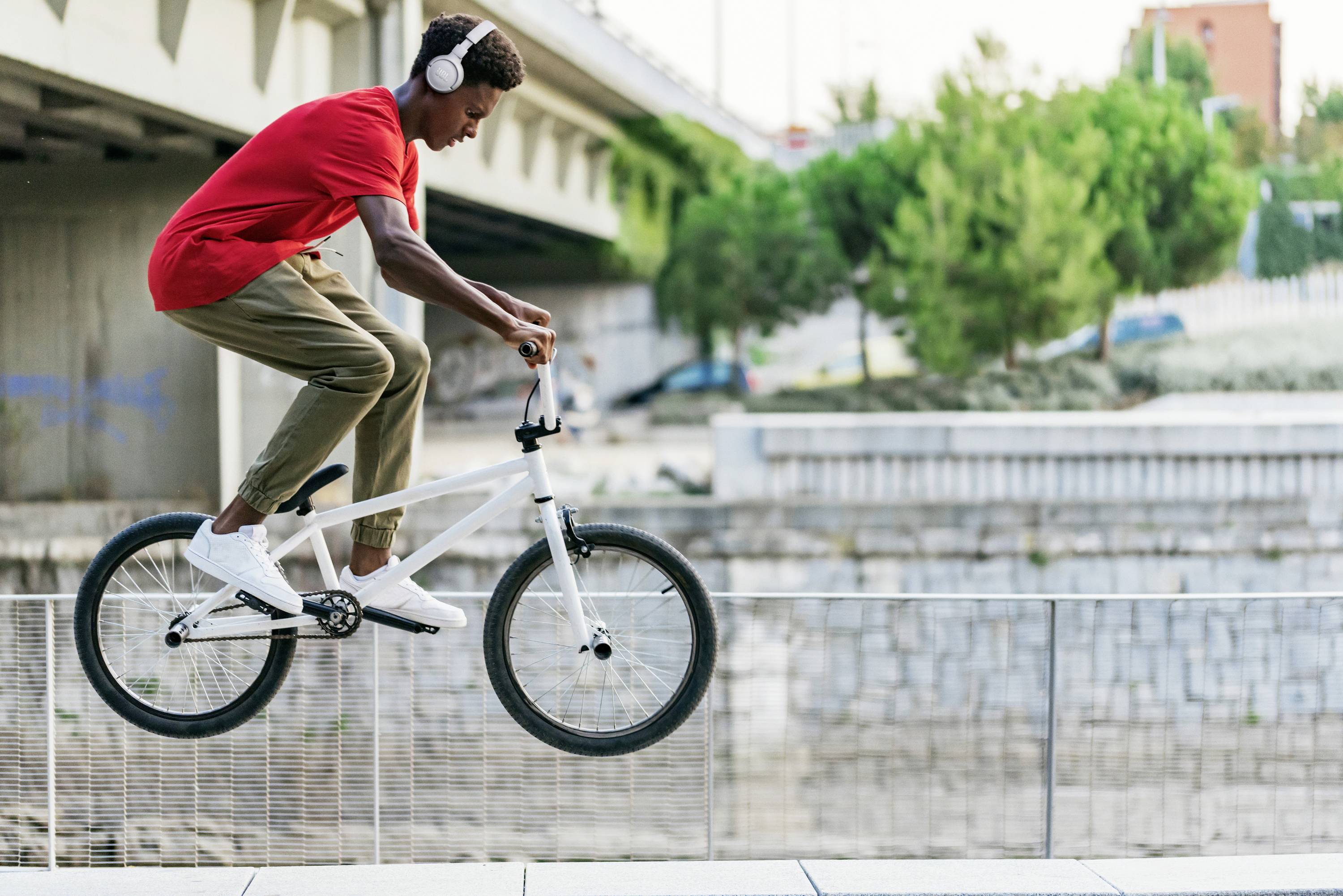 Ein junger Mann in rotem T-Shirt und Kopfhörern fährt mit einem BMX-Rad auf einem Geländer entlang. Im Hintergrund sind Bäume und eine Brücke.