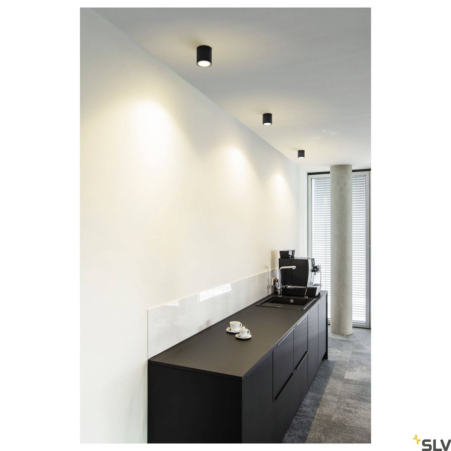 Schwarze Küchenzeile mit Kaffeemaschine und Tassen auf Arbeitsplatte. Oben sind drei zylindrische Deckenleuchten angebracht.