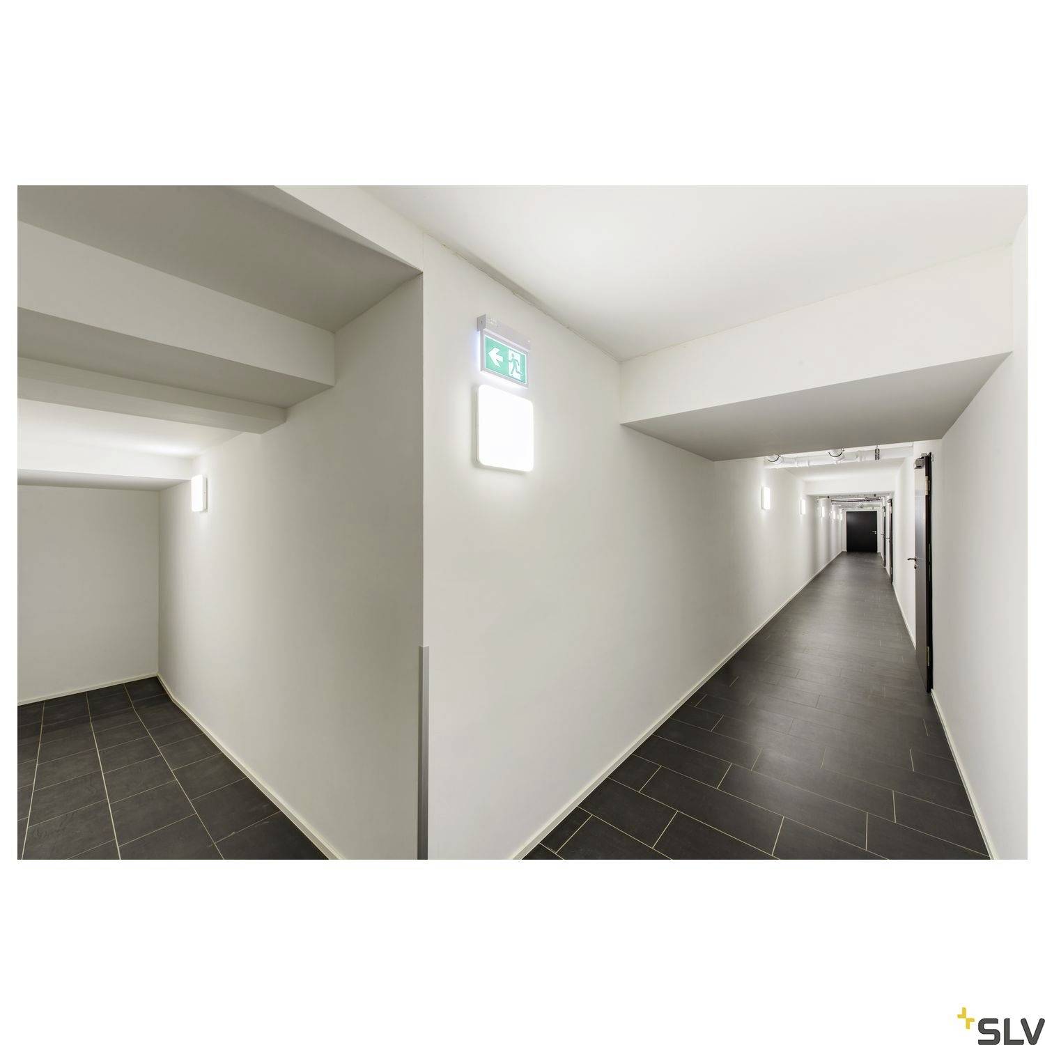 Ein langer, heller Flur mit schwarzen Fliesen und weißen Wänden. An der Decke hängen Leuchten, und ein Notausgangsschild leuchtet an der Wand.