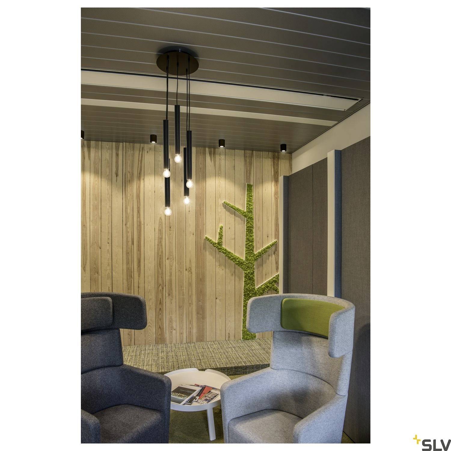 Moderner Raum mit Holzwand und Moosdeko. Zwei graue Sessel stehen um einen kleinen Tisch mit Zeitschriften. Hängende Glühlampen beleuchten den Bereich.