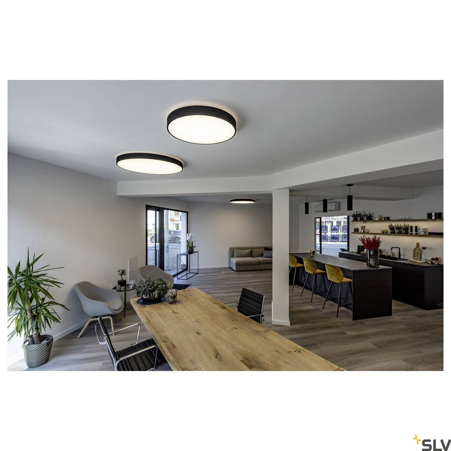 Ein modern eingerichtetes Wohnzimmer mit einem großen Holztisch, Pflanzen, einer grauen Couch und mehreren runden Deckenleuchten.