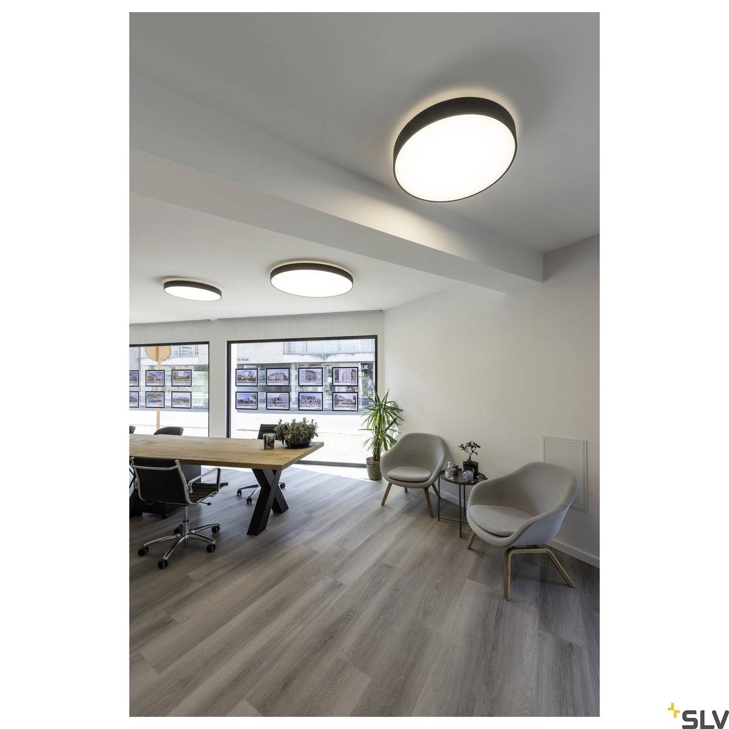 Konferenzraum mit hellem Holztisch, schwarzen Bürostühlen und zwei Sesseln. Große runde Deckenleuchten sorgen für Beleuchtung.