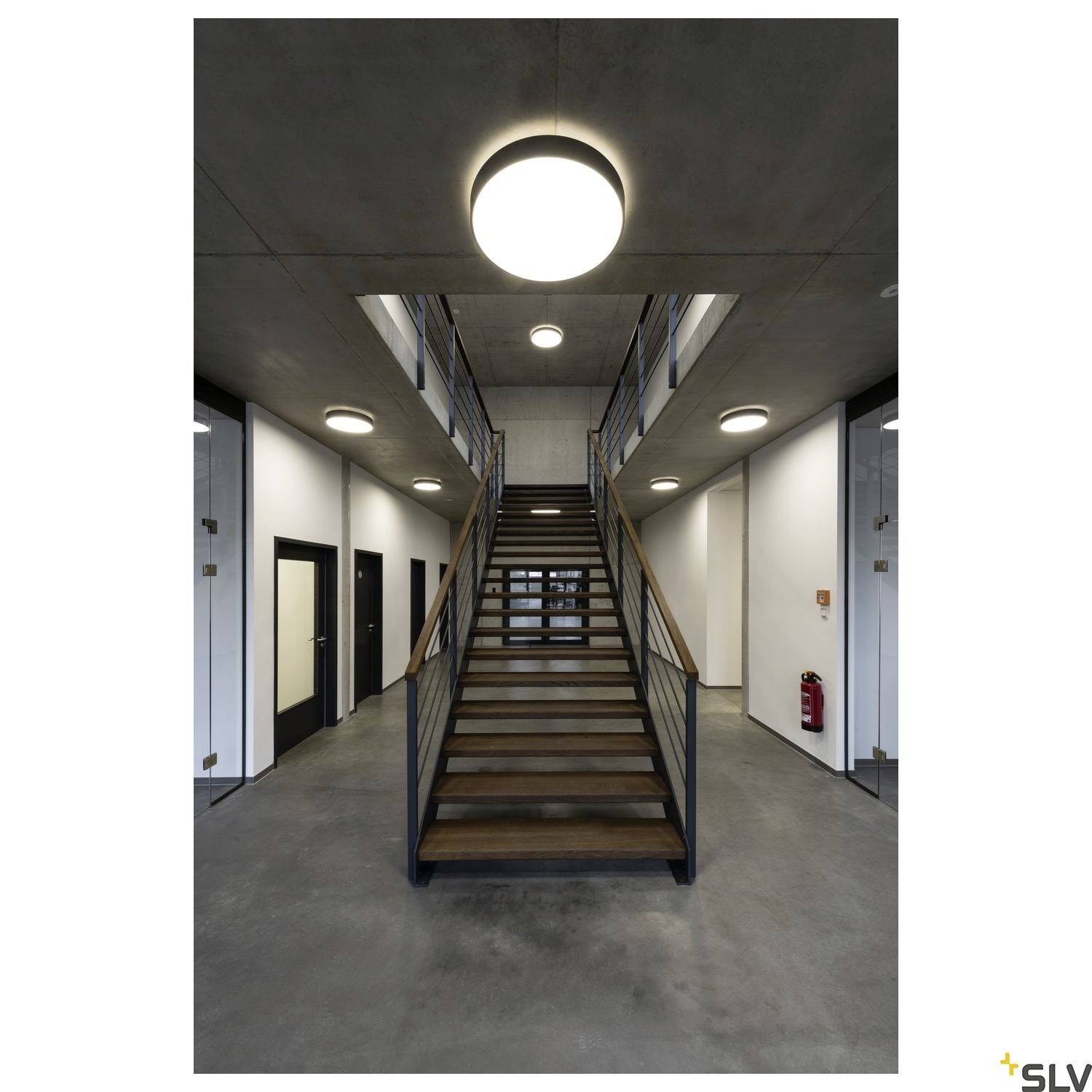 Eine moderne Treppe in einem Innenraum mit Betonwänden, Metallgeländern und runden Deckenleuchten. Der Boden ist grau.