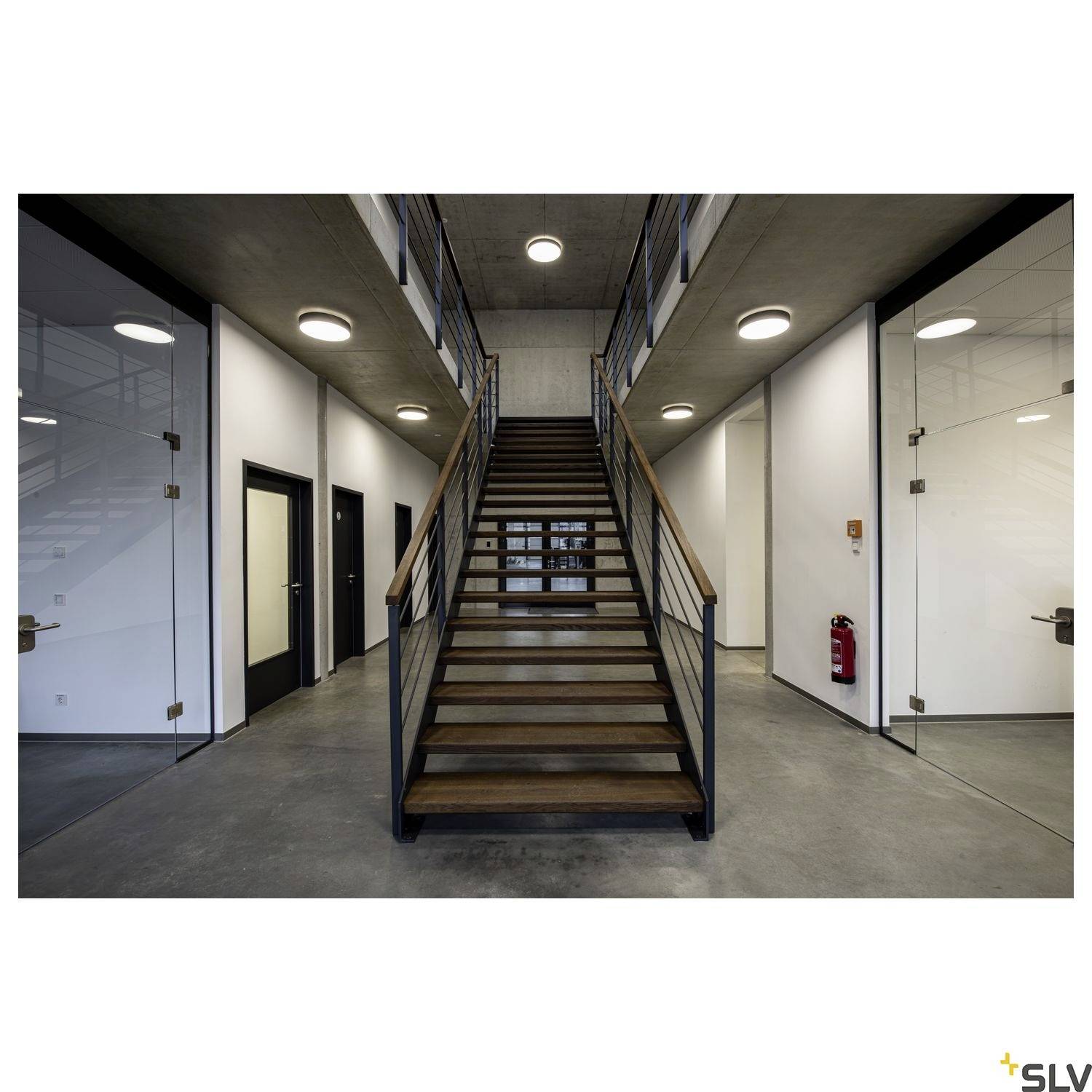 Ein modernes Bürogebäude mit einer zentralen Holztreppe, umliegenden Glaswänden und dunklen Türen, unter moderner Deckenbeleuchtung.