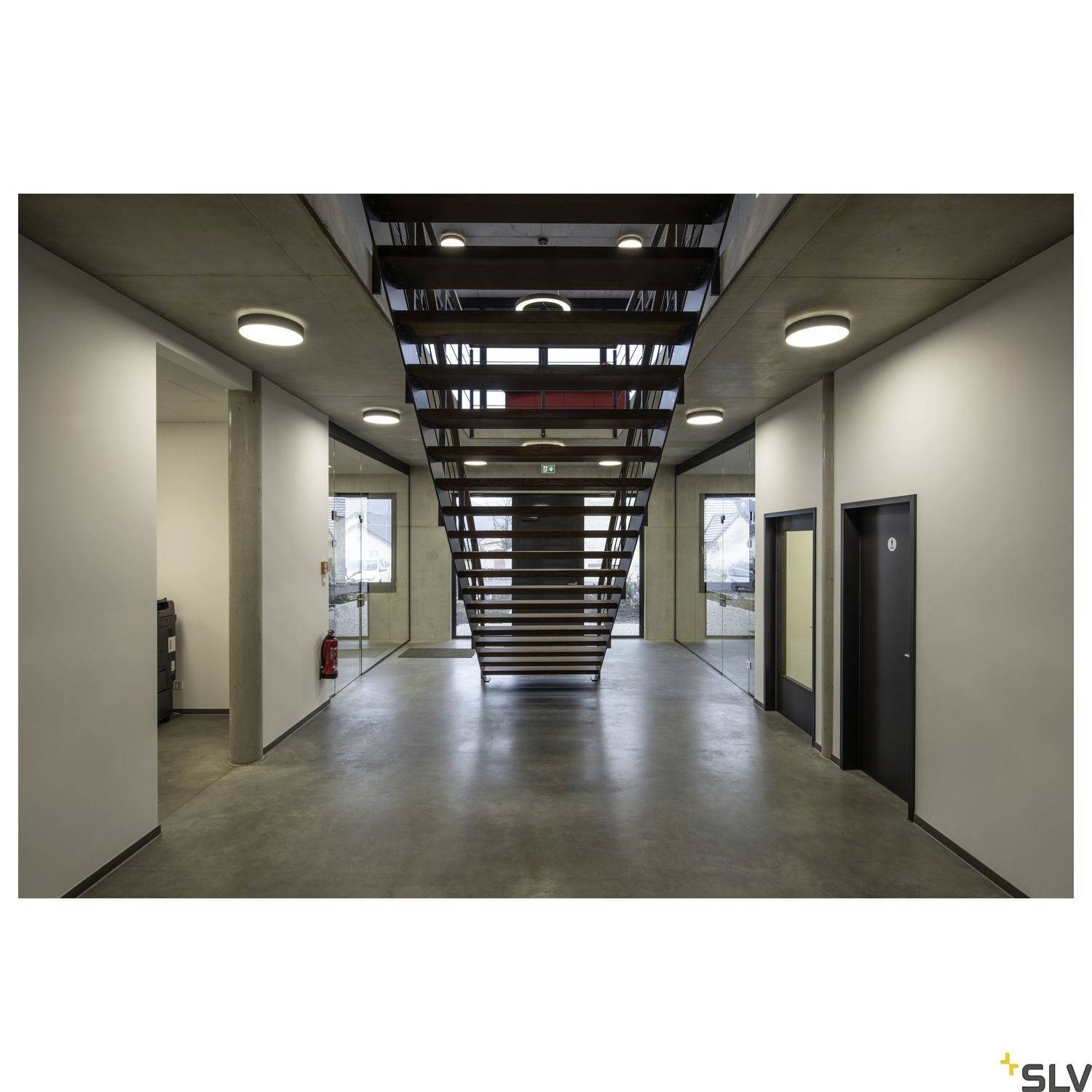 Ein moderner Innenraum mit einer zentralen Treppe, umgeben von weißen Wänden und Deckenleuchten. Links ein Feuerlöscher.