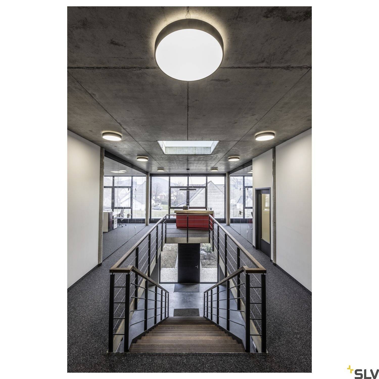 Offenes Bürogebäude mit zentralem Treppenaufgang, modernen Deckenleuchten und großen Fenstern, die natürliches Licht hereinlassen.