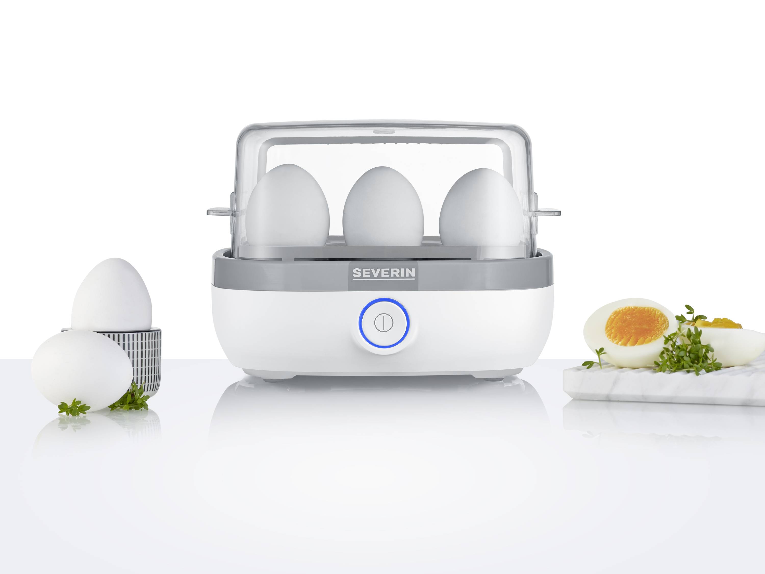 Ein Eierkocher mit vier Eiern im Gerät und einem gekochten, halbierten Ei auf einem Brett. Der Eierkocher ist weiß mit grauen Akzenten.