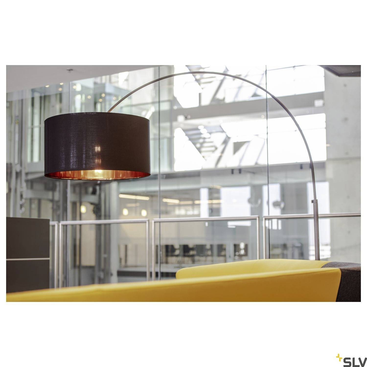 Hängelampe mit gebogenem Ständer leuchtet über einem gelben Sofa in einem modernen Bürogebäude mit Glaswänden und metallischen Strukturen.