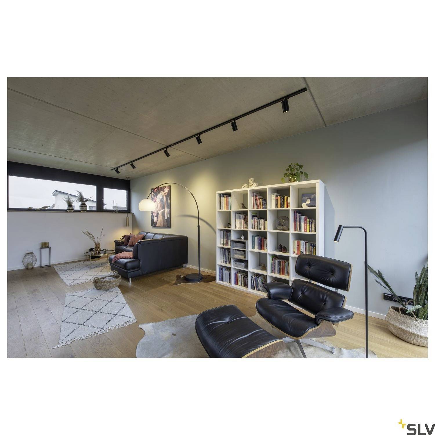 Wohnzimmer mit Parkettboden, moderner schwarzer Sessel, Bücherregal, Stehlampe und großen Fenstern mit Blick auf die Stadt.