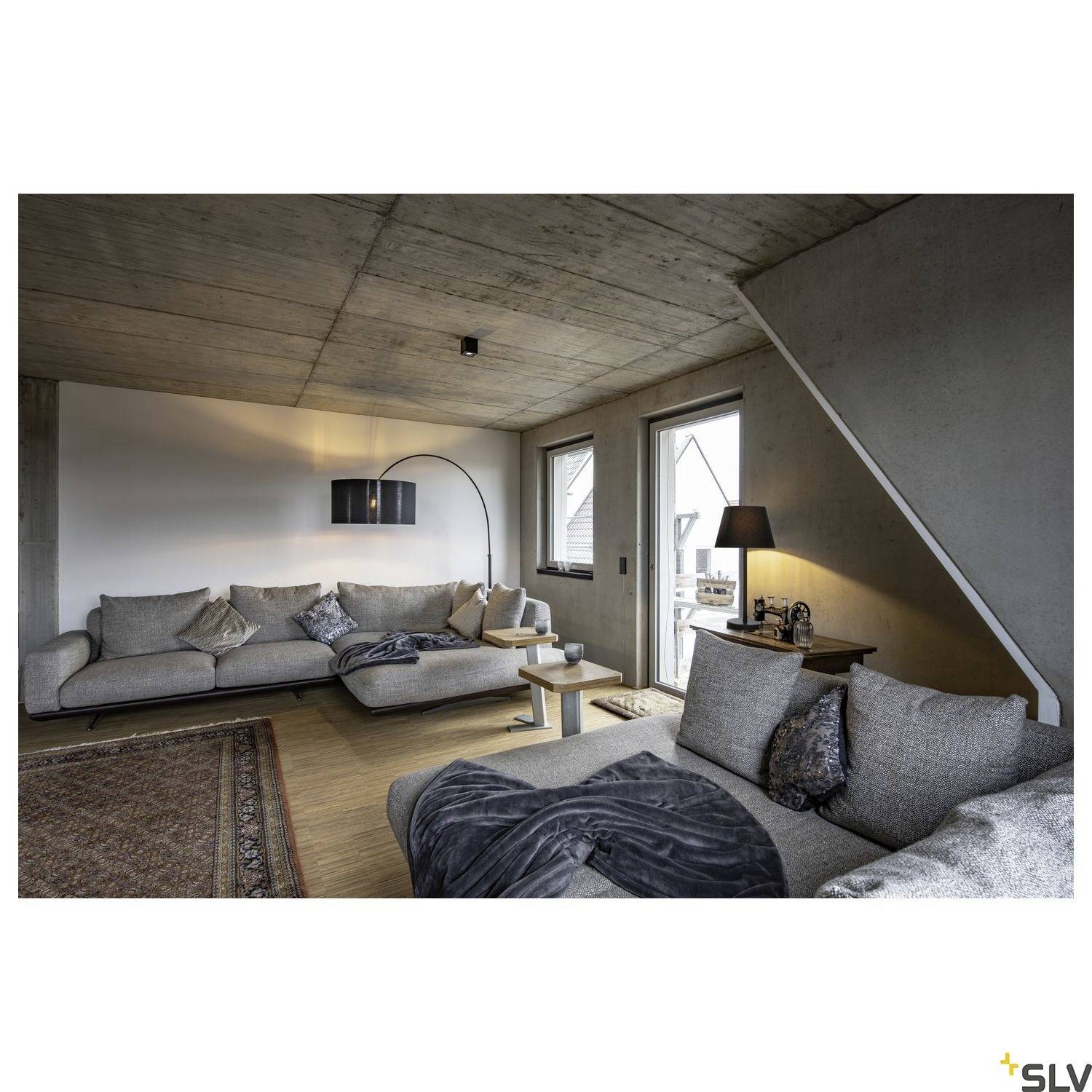 Gemütliches Wohnzimmer mit grauem Sofa, Stehlampe, Couchtisch und Teppich auf Holzboden, Beton- und Holzwände, Tageslicht durch Fenster.