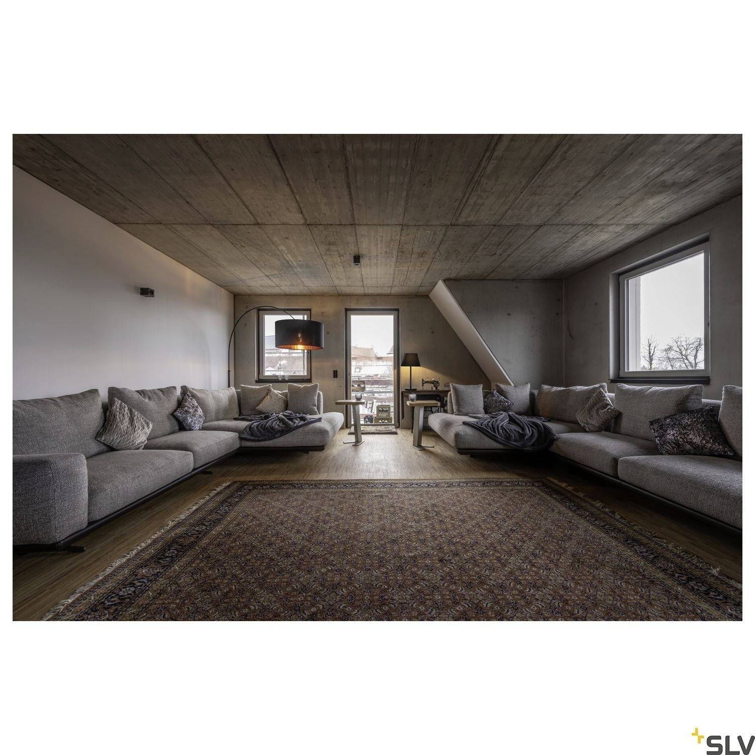 Großes Wohnzimmer mit zwei grauen Sofas, vielen Kissen und einem Teppich in der Mitte. Große Fenster lassen viel Tageslicht herein.