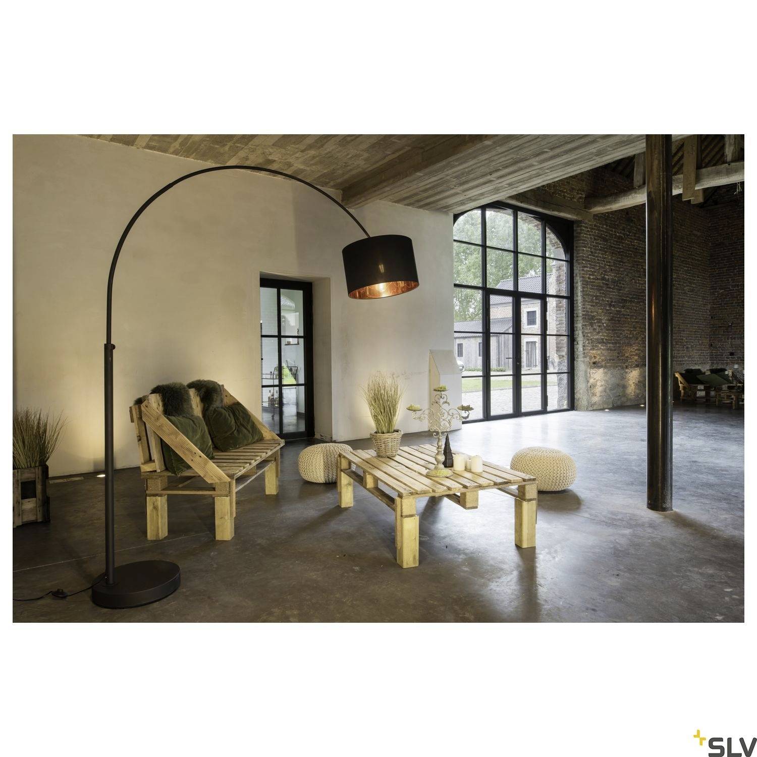 Gemütlicher Wohnbereich mit gebogenen Stehlampe, Holzsofa mit grünen Kissen, Palettentisch und großen Fenstern in einem Loft-Stil-Raum.
