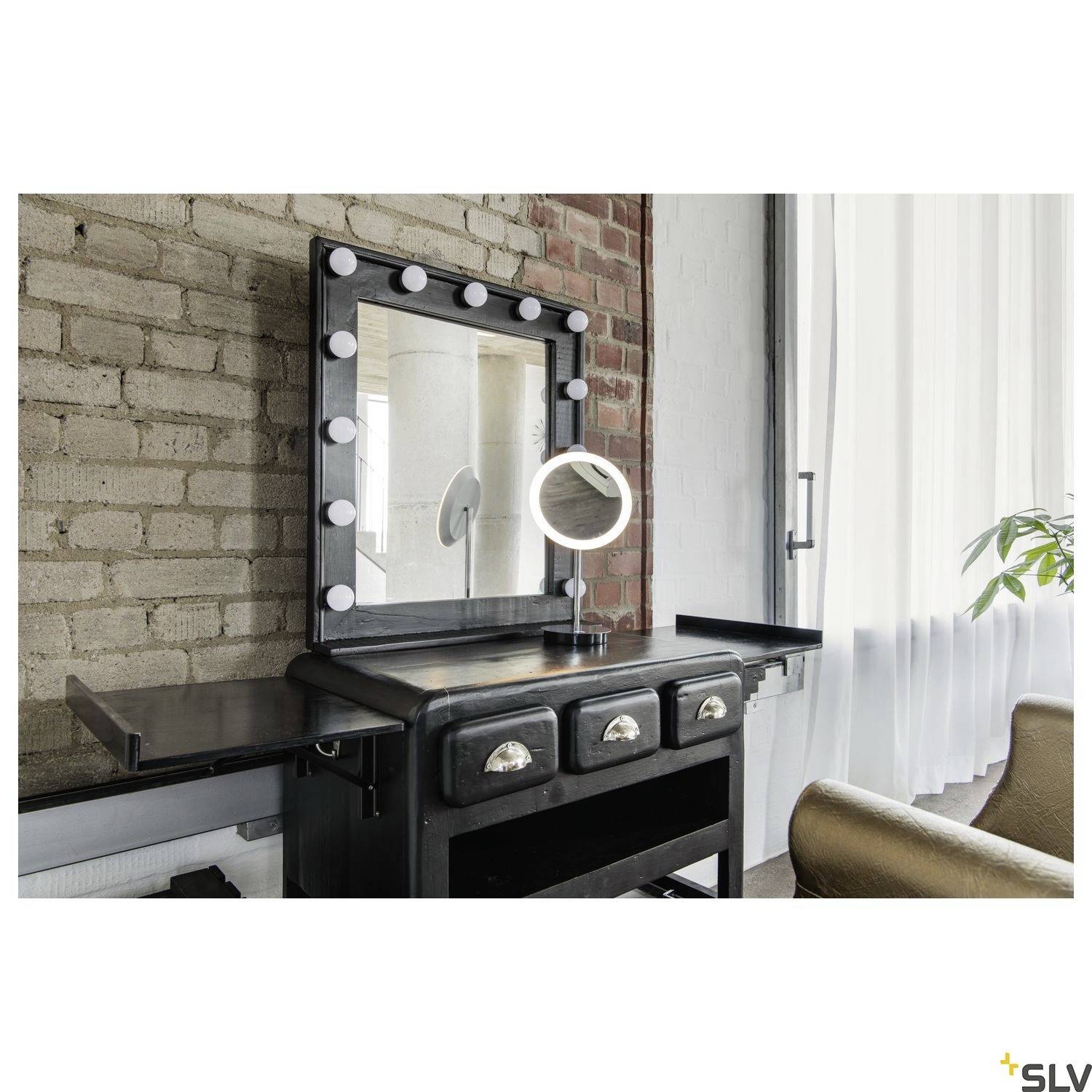 Frisiertisch mit beleuchtetem Spiegel vor einer Backsteinwand, daneben ein brauner Stuhl und ein Fenster mit weißem Vorhang.