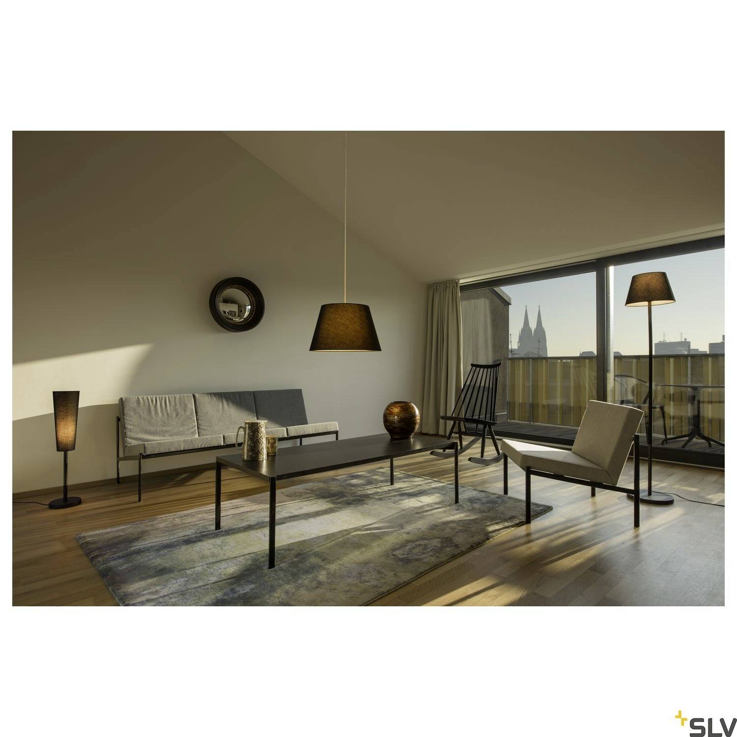 Ein modernes Wohnzimmer mit Sofa, Tisch und Stehlampen. Große Fenster bieten Blick auf den Dom im Hintergrund.