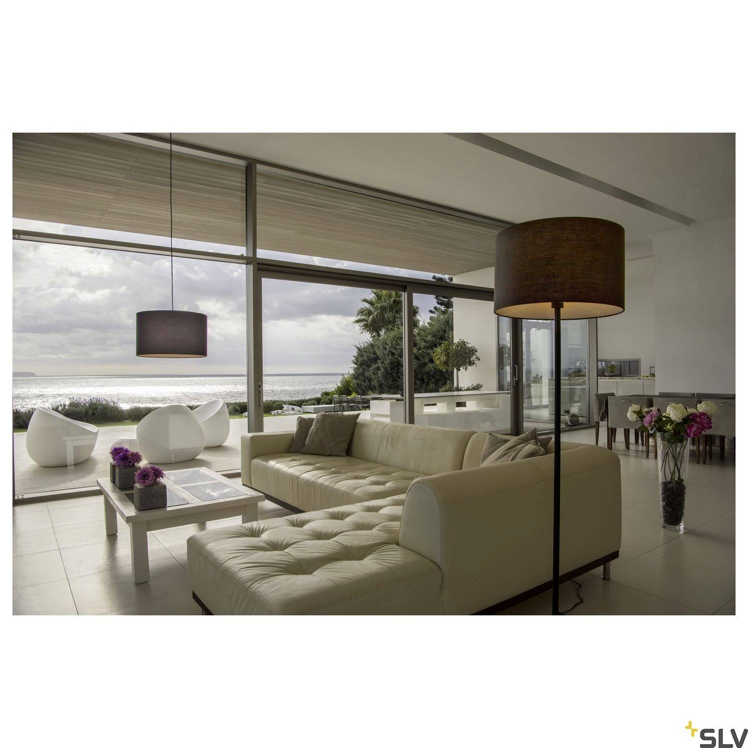 Modernes Wohnzimmer mit großen Fenstern zum Meer, Sitzgruppe, Stehlampe und dekorativem Tisch, ruhige und elegante Atmosphäre.