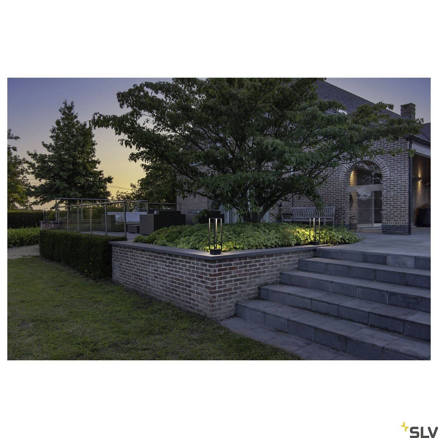 Das Bild zeigt einen gepflegten Garten mit beleuchteten Stufen und einem großen Baum vor einem Backsteinhaus in der Abenddämmerung.