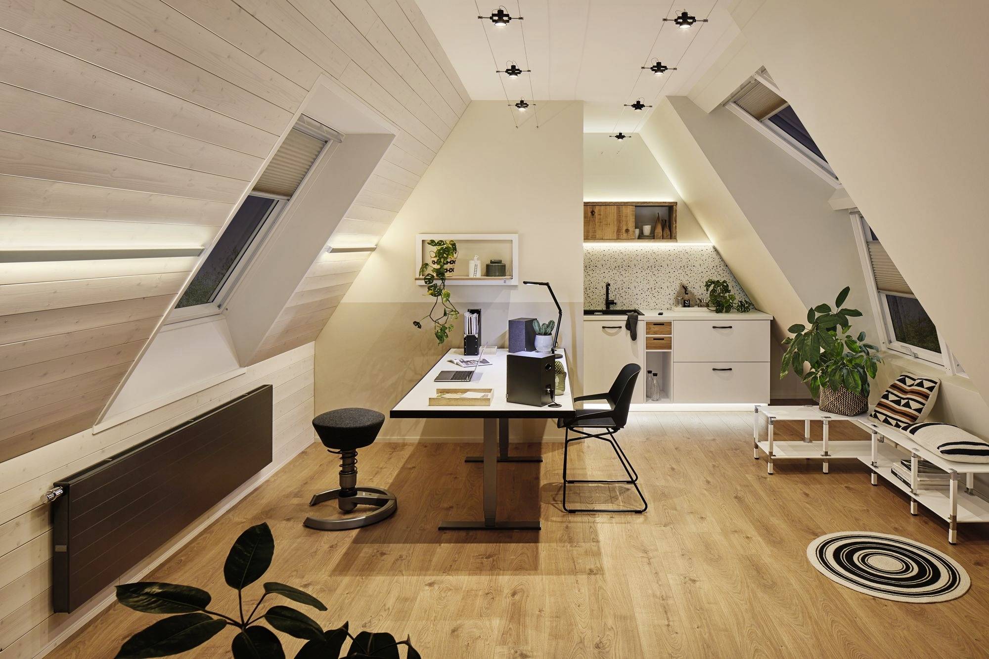 Dachgeschossbüro mit Schreibtisch, Laptop, Stuhl und Hockern. Moderne integrierte Küche im Hintergrund. Helle Holzverkleidung und Beleuchtung.