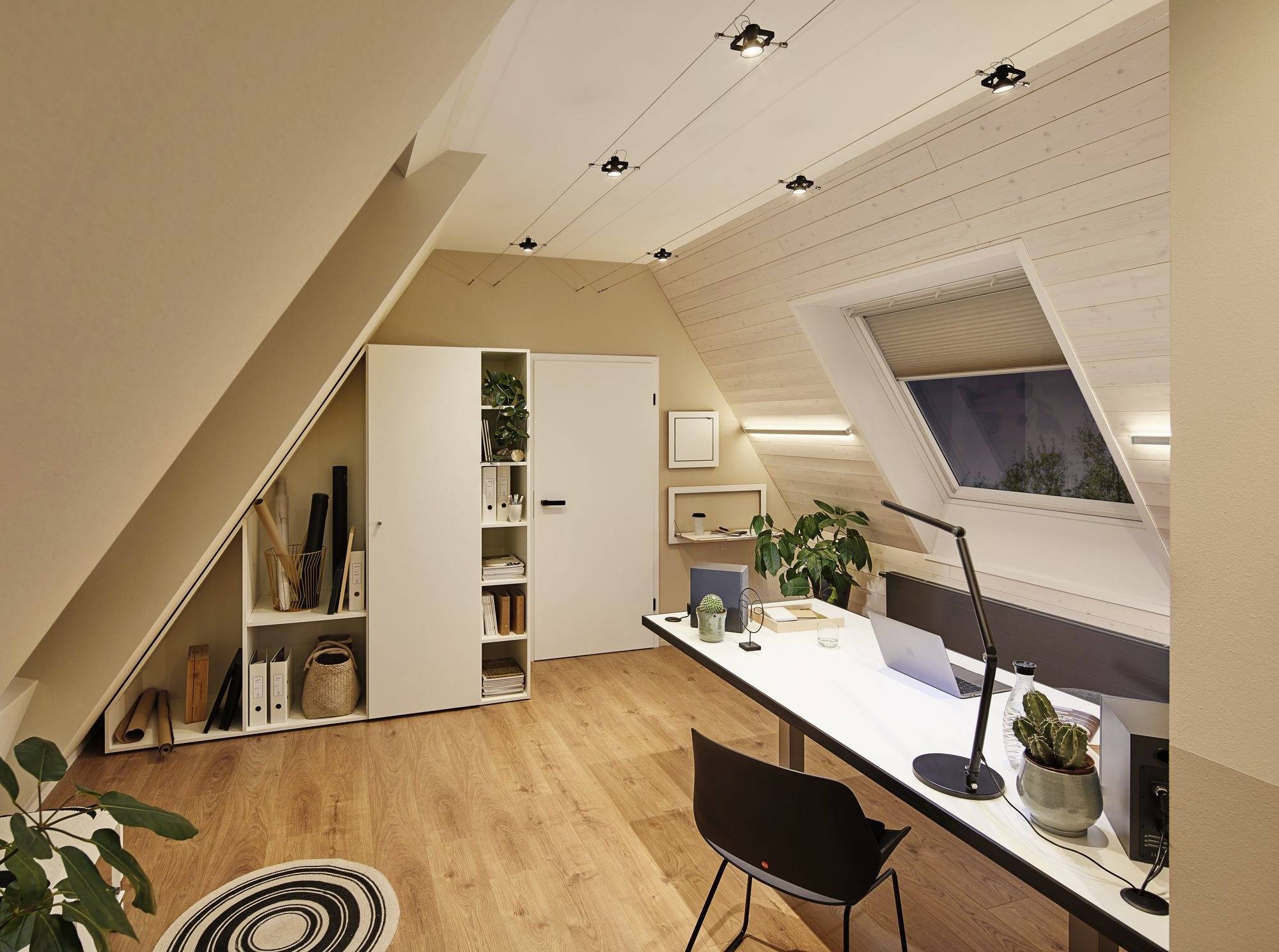 Ein modernes, gut beleuchtetes Dachgeschossbüro mit Holzfußboden. Ein langer Schreibtisch mit Laptop und Pflanzen. Regal mit Ordnern.