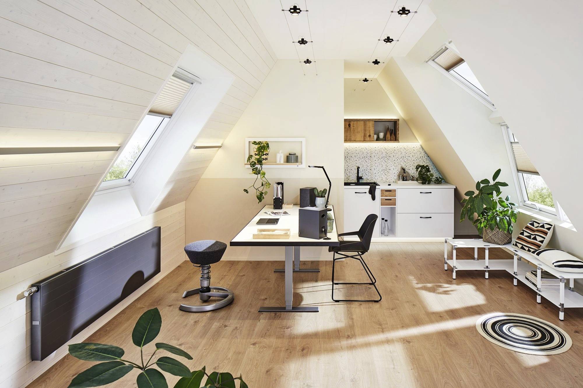 Ein modernes, lichtdurchflutetes Dachgeschossbüro mit Schreibtisch, Stuhl, Pflanzen und einer kleinen Küchenzeile im Hintergrund.