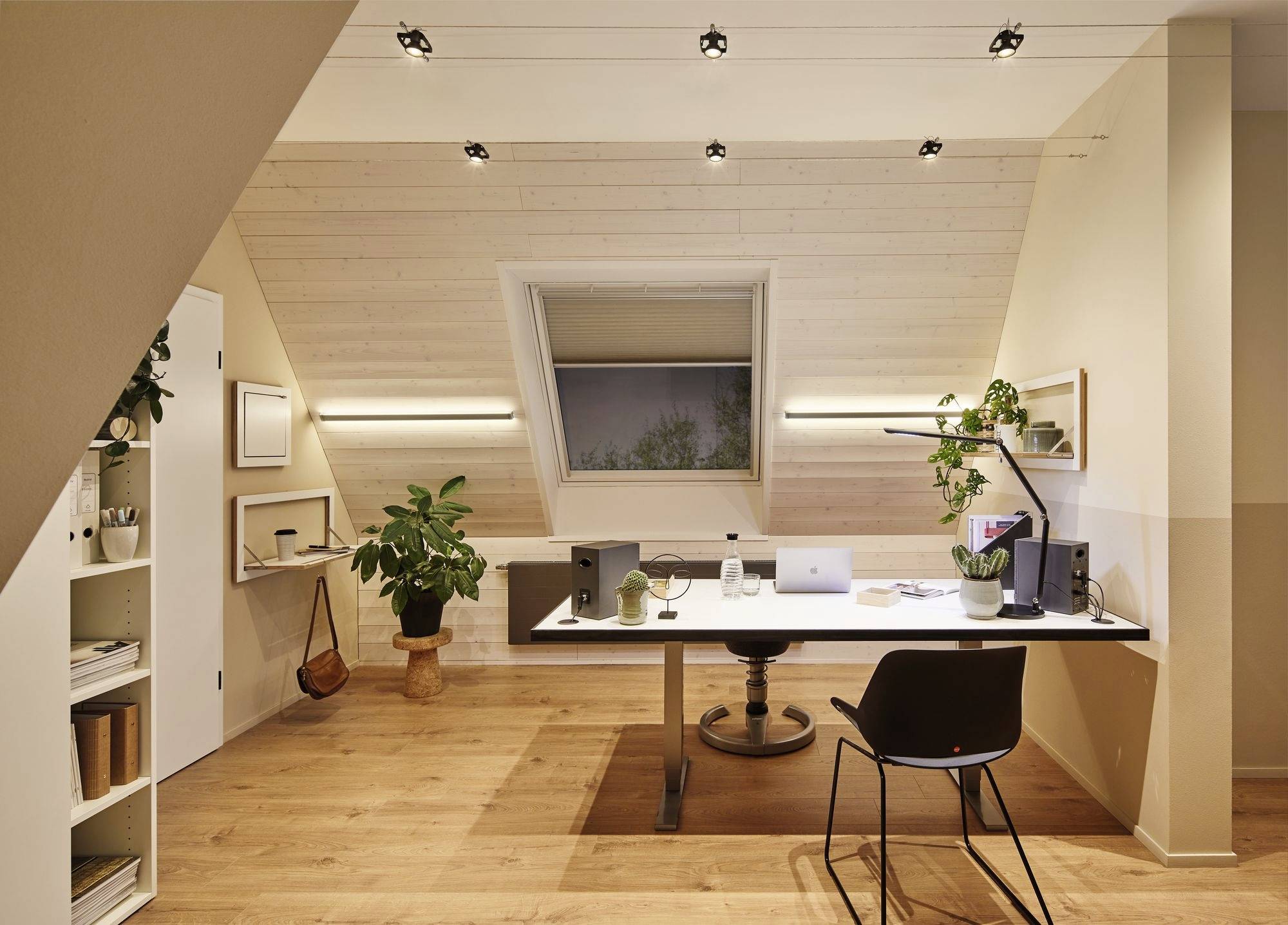 Ein modernes Arbeitszimmer mit schräger Decke, großem Schreibtisch, Laptop, Pflanzen und Regalen. Warmes Licht und Holzfußboden.