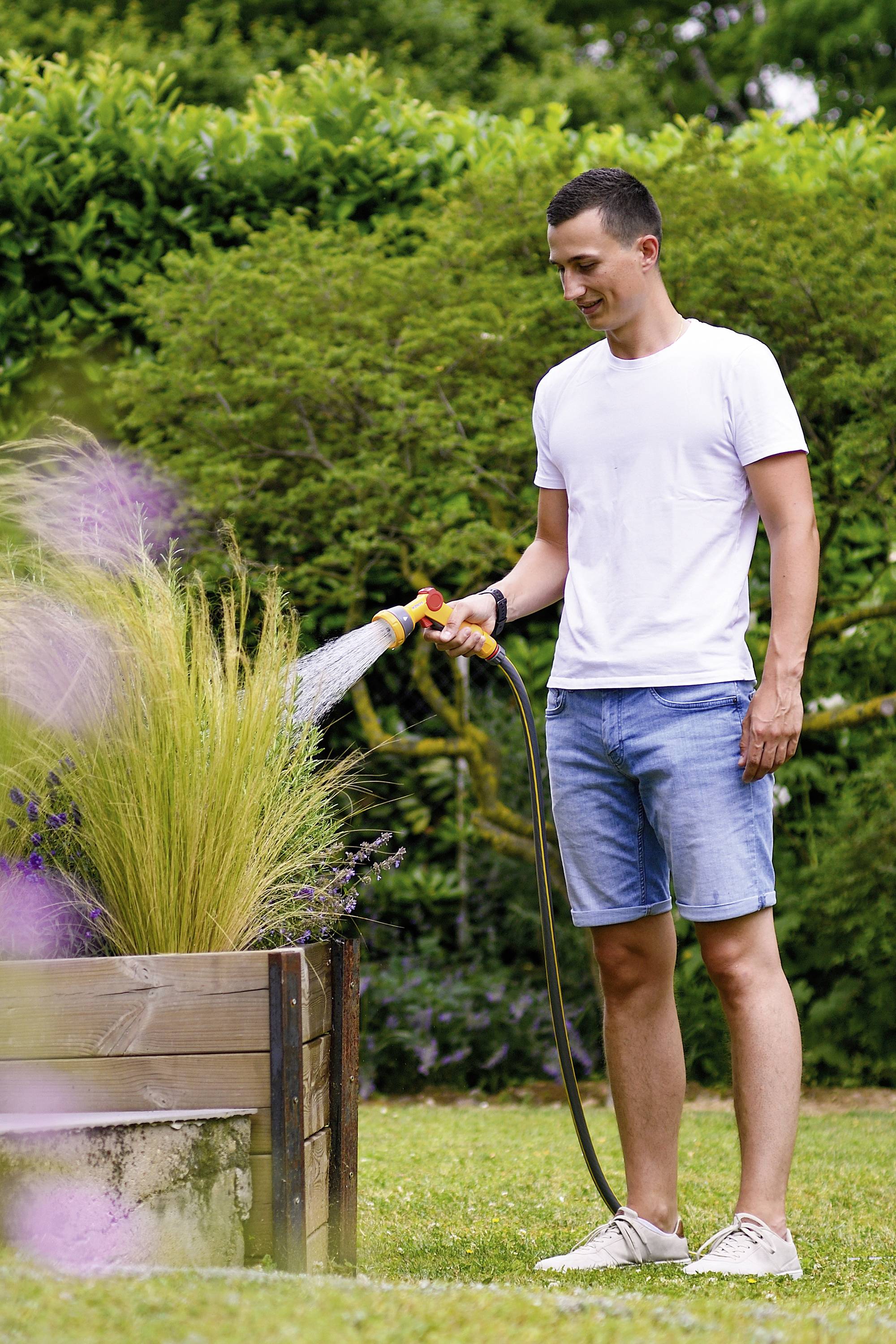Ein Mann im weißen T-Shirt und Jeansshorts gießt Pflanzen in einem Holzbeet mit einem Gartenschlauch in einem grünen Garten.