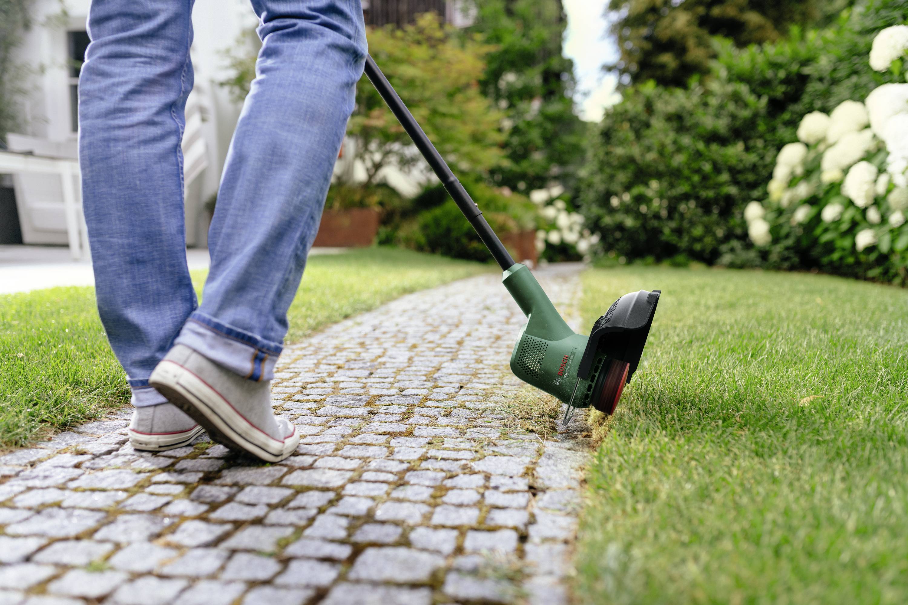 Eine Person schneidet mit einem elektrischen Rasentrimmer Gras entlang eines gepflasterten Weges in einem Garten.