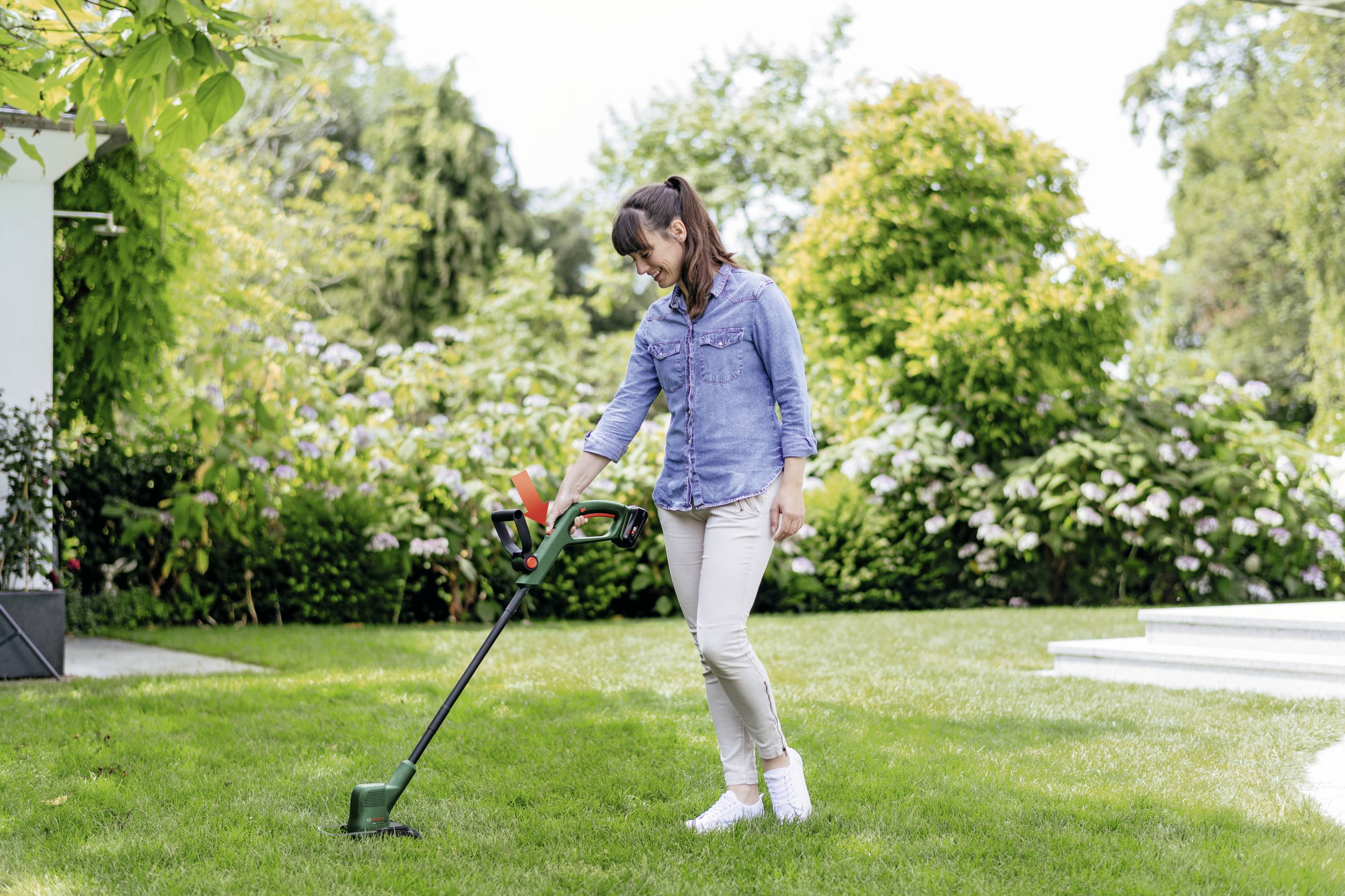 Frau mäht mit einem elektrischen Rasentrimmer Rasen im Garten; im Hintergrund grüne Bäume und Büsche.