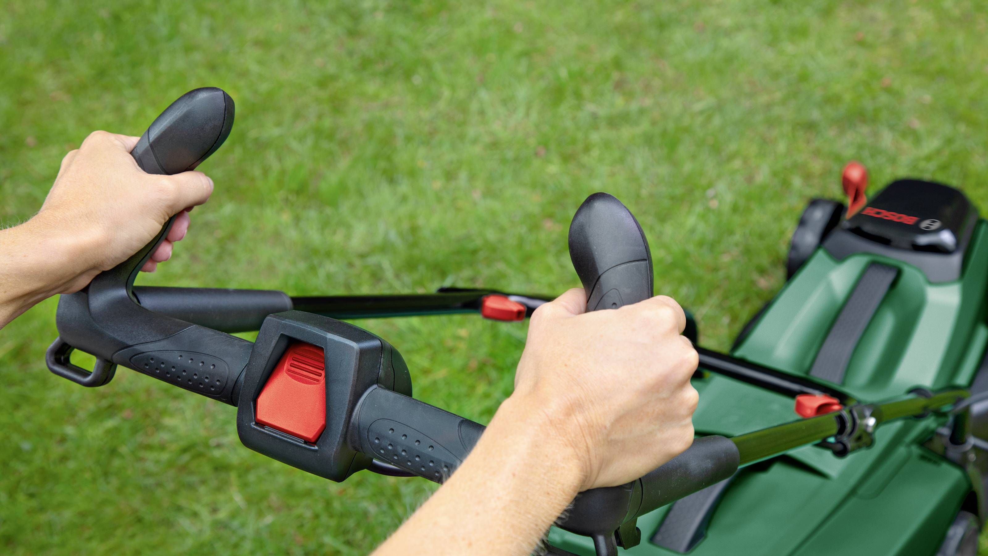Eine Person hält den Griff eines Rasenmähers auf grünem Gras. Die Hände umfassen zwei ergonomische Griffe zur Steuerung des Geräts.