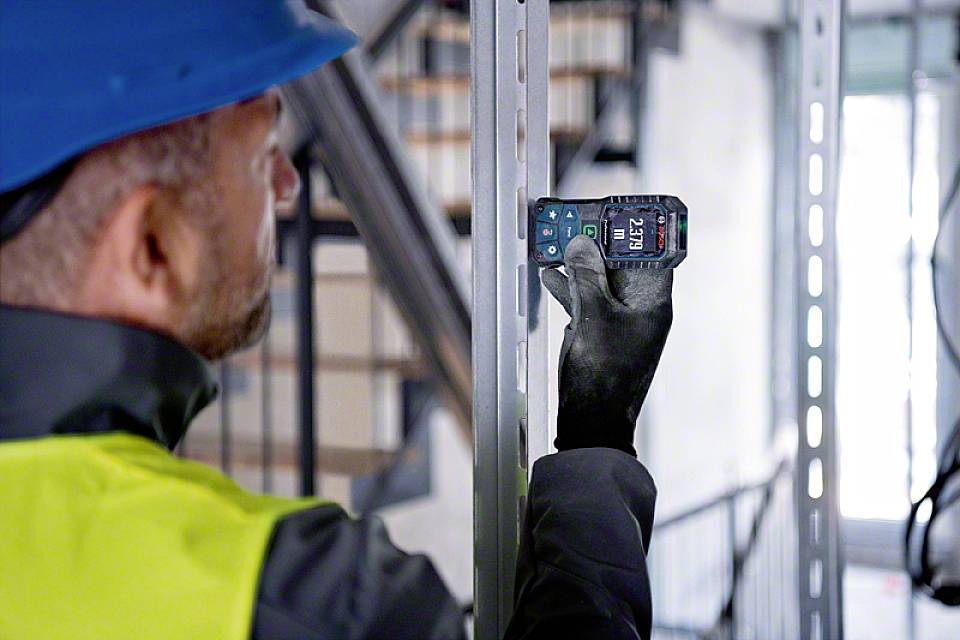 Ein Bauarbeiter mit Schutzhelm und Weste misst mit einem Entfernungsmesser die Distanz in einer industriellen Umgebung.