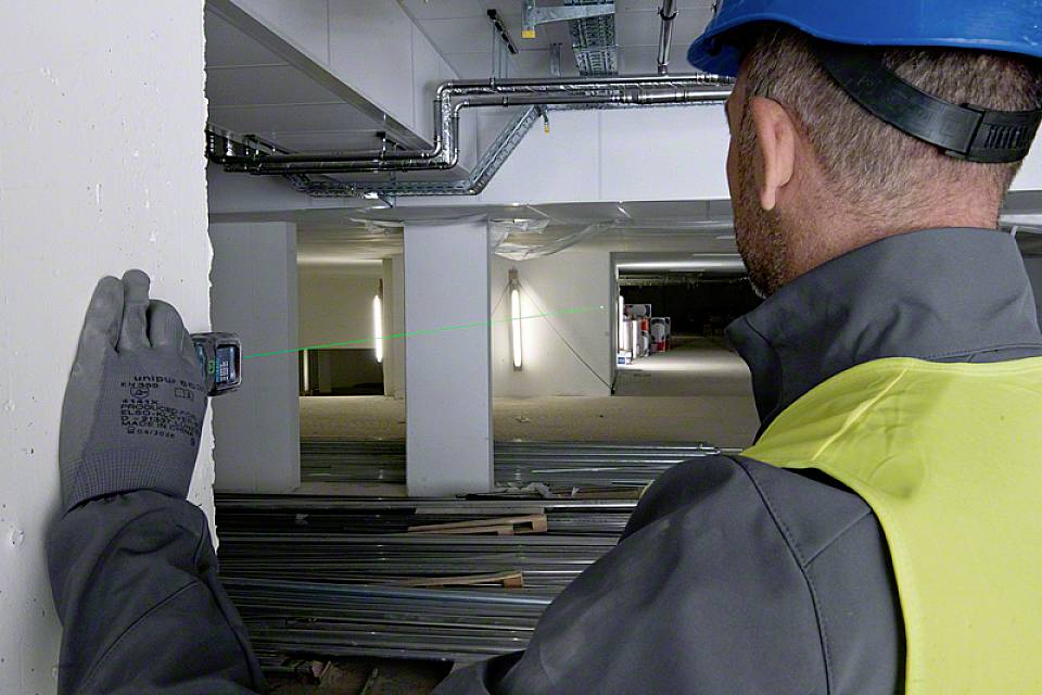 Ein Bauarbeiter in Schutzkleidung misst mit einem Lasergerät die Entfernung in einem unfertigen Gebäude. Baustellenumgebung mit Metallträgern.