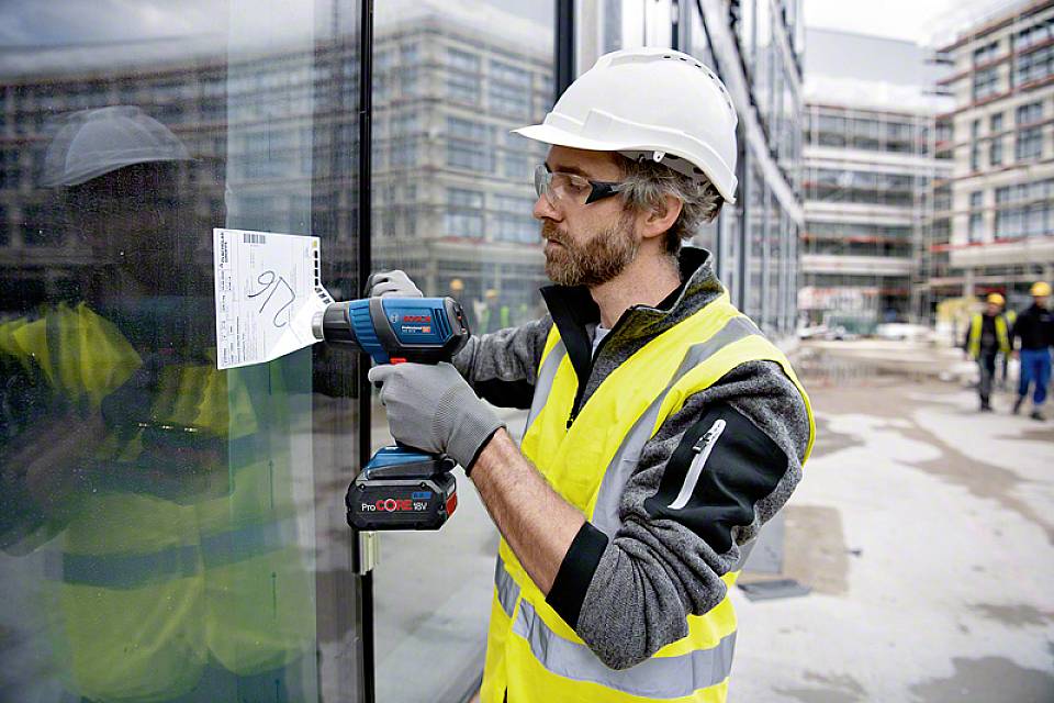 Ein Bauarbeiter mit Schutzhelm und Sicherheitsweste befestigt mit einem Akkuschrauber ein Dokument an eine Glaswand auf einer Baustelle.