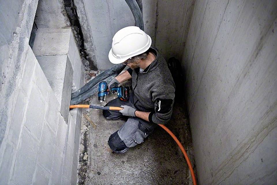 Ein Arbeiter in Schutzkleidung und Helm befestigt ein orangefarbenes Kabel an einer Betonwand mit einem Elektrowerkzeug.